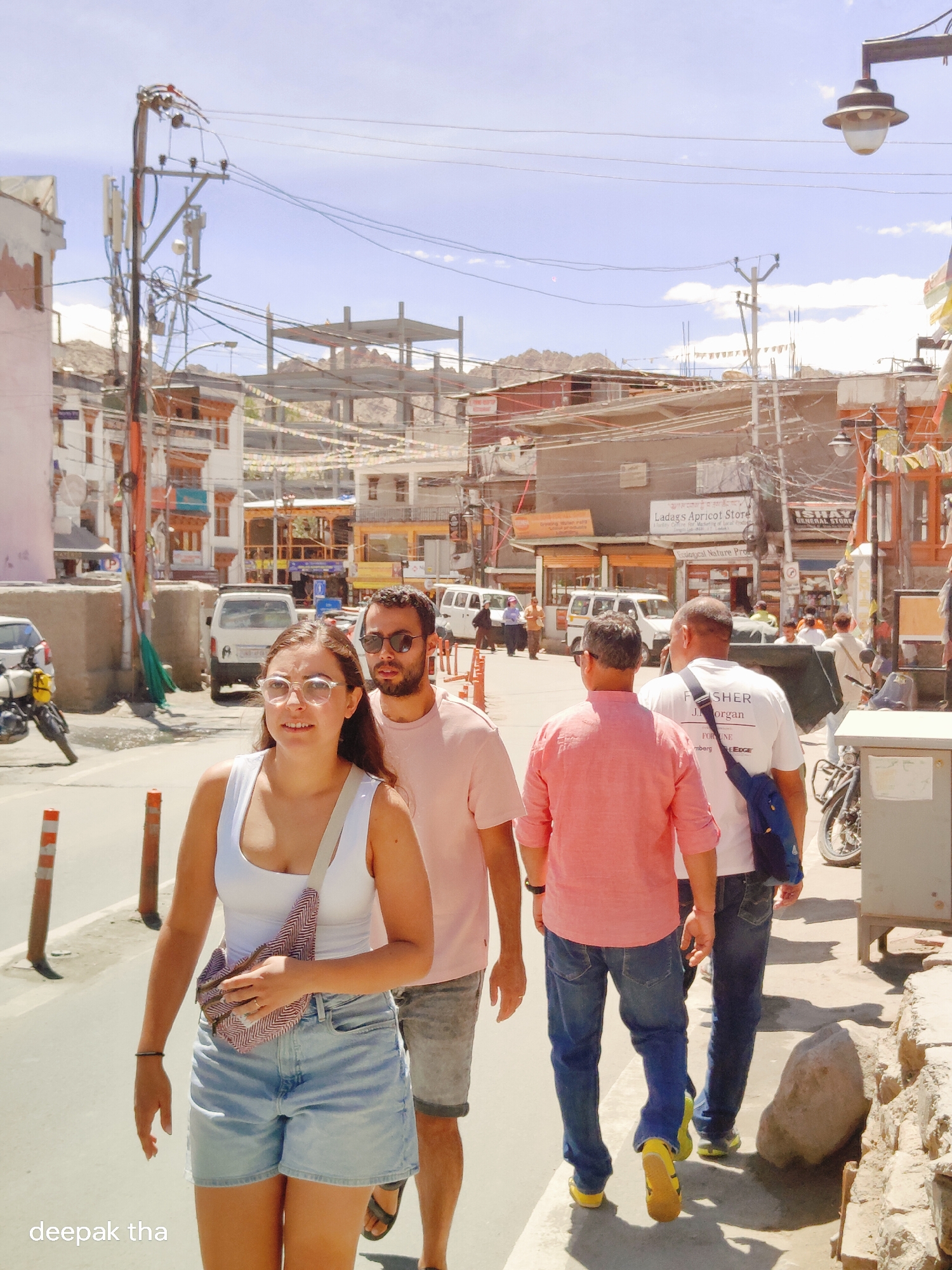 The image captures a street scene in Leh, India, under a bright, partly cloudy sky. A group of four individuals walks along a paved sidewalk on the right side of the image, with a paved road on the left. In the foreground, a woman with glasses and a white tank top, wearing denim shorts and a patterned fanny pack, walks ahead. She is followed by three men. The first man behind her, wearing a pink t-shirt and shorts, is partially obscured by her. Further back, two men in casual wear, one in a pink shirt and jeans, and the other in a white t-shirt and jeans with a blue bag slung over his shoulder, are walking away from the camera.
The environment is characteristic of a bustling Indian town, with buildings lining both sides of the street. These buildings appear to be a mix of residential and commercial structures, with shops displaying signage, including "Ladakh Apricot Store" and "Fisher General Store." Overhead, a complex network of power lines and utility poles crisscrosses the sky, with festive bunting strung between some of the buildings. In the background, unfinished construction of a multi-story building is visible, and further in the distance, mountains are silhouetted against the sky. A few vehicles, including cars and a motorcycle, are present on the road. The overall impression is a snapshot of daily life in Leh, with a blend of traditional architecture and modern infrastructure.