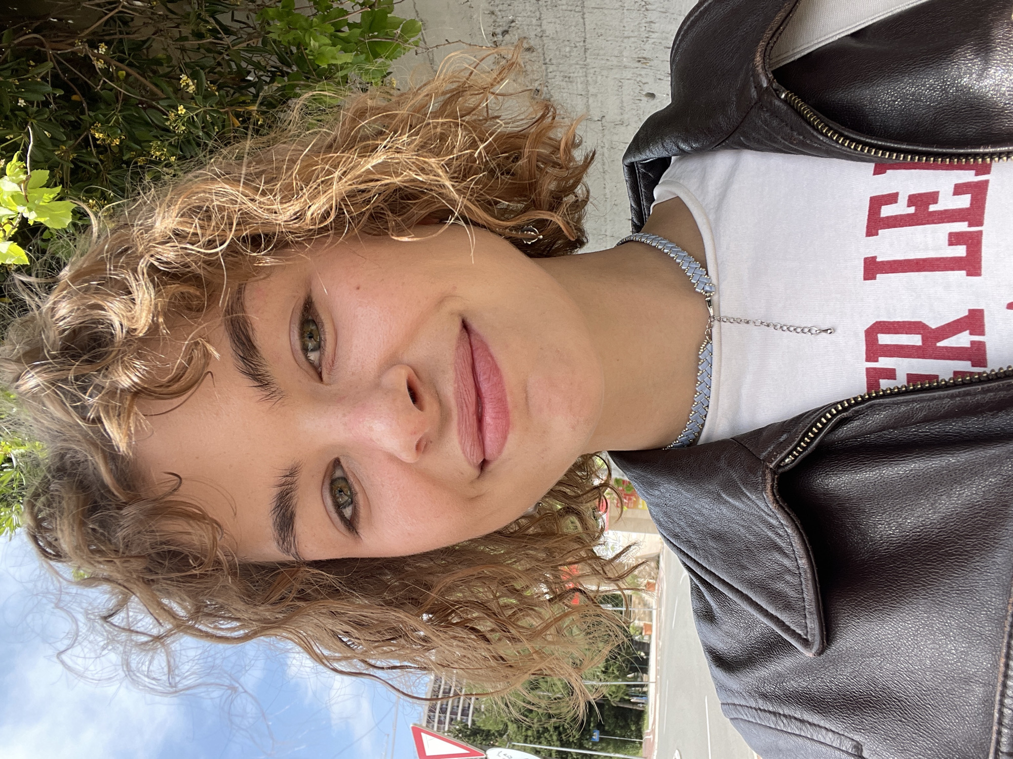 The image is a selfie of a woman with curly light brown hair, wearing a white t-shirt with red lettering partially obscured by a dark brown leather jacket. She has light skin, green eyes, and a light pink lip color. She is also wearing a blue and silver beaded necklace. The background includes greenery, suggesting she is outdoors. A building and street sign are partially visible in the background, indicating an urban environment. The photo was taken in Rafailovići, Montenegro, as indicated by the street sign's typography and design, along with the style of the architecture.