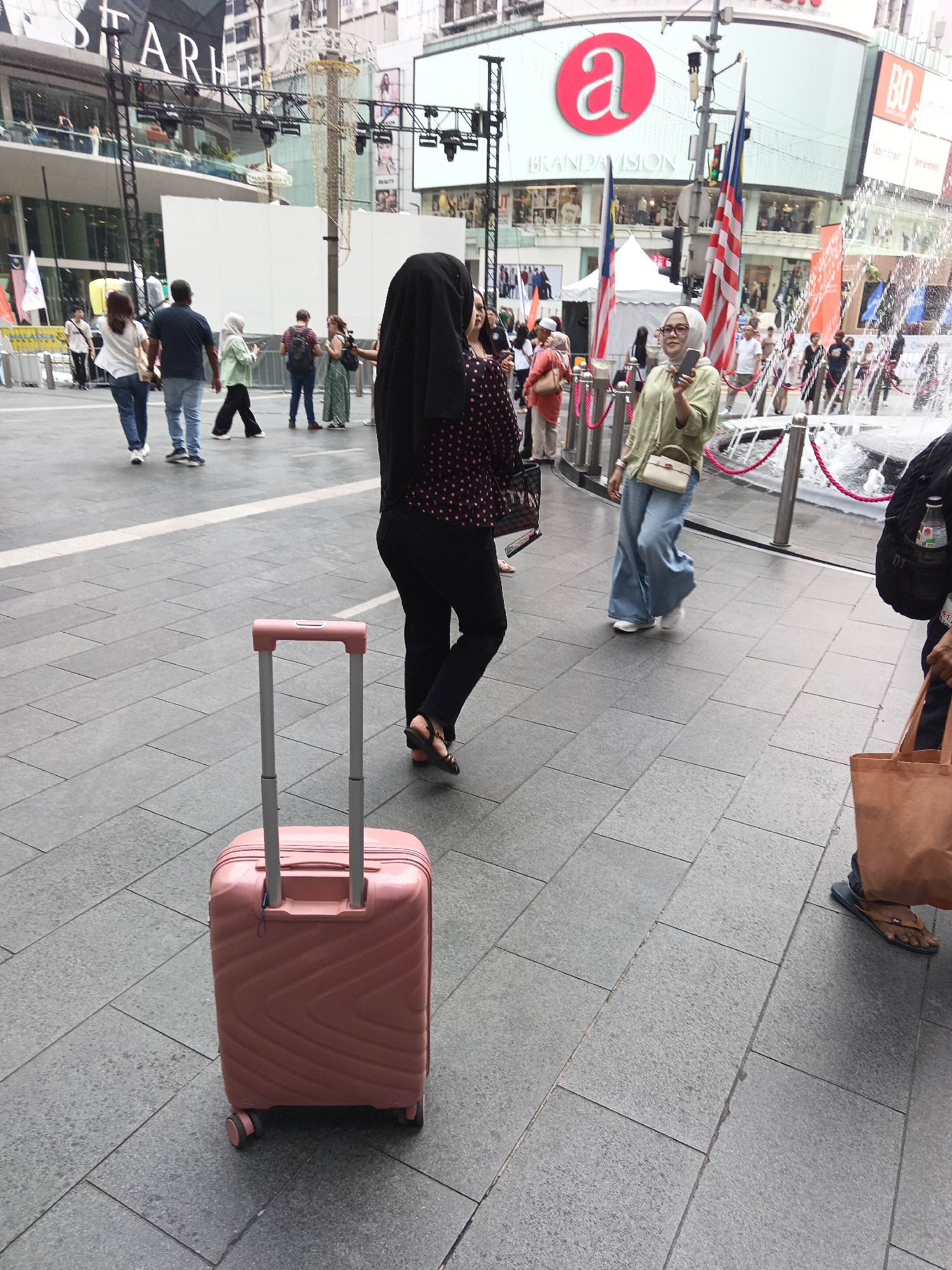 The image depicts a street scene in Kuala Lumpur, Malaysia, likely near a shopping area given the presence of commercial buildings in the background with prominent signage. In the foreground, a light pink suitcase with a telescoping handle stands on a grey tiled pavement. To the right of the suitcase, a person wearing black pants and a patterned top, with their back to the camera, is walking away. Further into the scene, several other individuals are walking in different directions. One person in the mid-ground is wearing a green top and blue wide-leg pants, holding a smartphone. Another person in the mid-ground is wearing a black headscarf and a black and pink polka dot top, holding a handbag. The background shows a large building with a prominent red "a" logo and "BRANDAVISION" text, suggesting it is a retail or commercial establishment. There are also flags, including the Malaysian flag, and a fountain with water spraying. The overall atmosphere appears to be a public urban space with pedestrian activity.