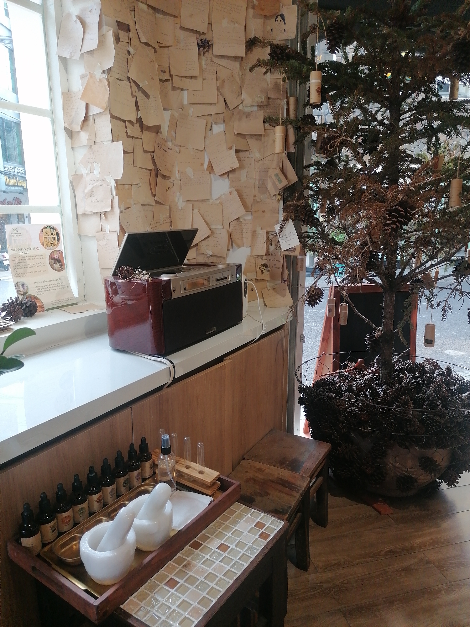 The image shows an interior space in Da Lat, Vietnam, possibly a shop or cafe, featuring a counter space. On the counter, there's a red and black electronic device, possibly a CD player or stereo system. Next to the device, there's a window with a view of the street outside. A poster with Vietnamese writing is placed near the window. The wall behind the counter is covered in numerous small pieces of paper or notes. Below the counter, there is a tray containing a mortar and pestle set, along with an assortment of small bottles, likely essential oils. A small wooden table is situated in front of the counter. To the right of the counter, a small tree, sparsely decorated with pinecones and hanging corks, sits in a pot filled with pinecones. The floor is made of wood. The scene is relatively still, with no apparent human interaction.