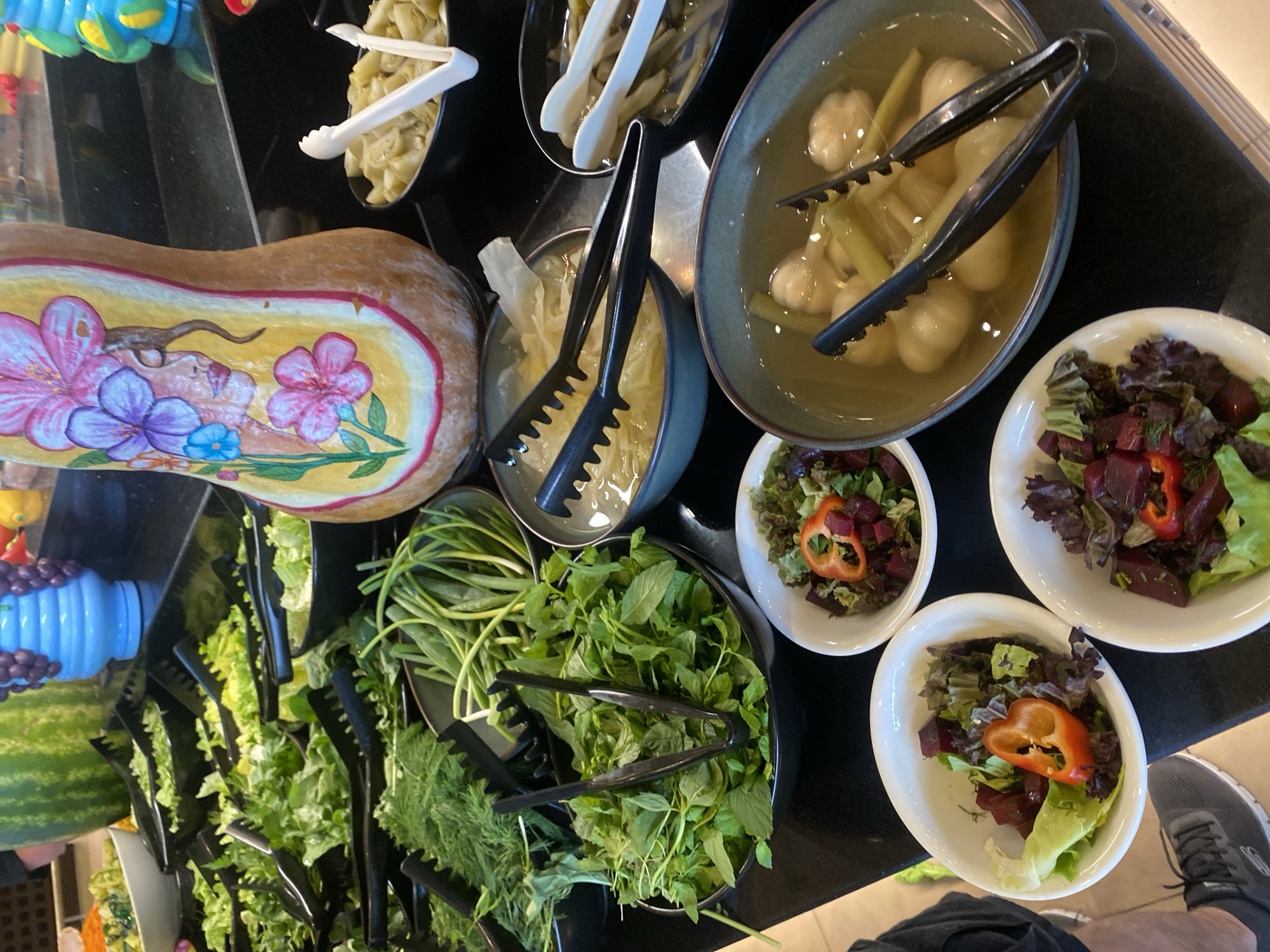 The image shows a buffet-style food spread. In the foreground, there are several white bowls containing salads made of lettuce, beets, and sliced red peppers. To the right, a large teal bowl holds pickled items, possibly garlic or small onions, in a light-colored brine, with black tongs resting inside. Further back and to the right, a black bowl contains what appears to be pickled vegetables, with white tongs inserted. To the left, a bowl holds fresh mint or similar herbs, with black tongs. Behind the herbs, there is a selection of green leafy vegetables and herbs in black serving dishes, some with tongs. In the background, a large watermelon is visible, as well as a blue decorative vase with purple grapes. A uniquely painted gourd, decorated with a floral design and a woman's face, stands prominently in the center of the frame. The food is arranged on a black countertop. The overall scene suggests a self-service food station. The location is not specified.