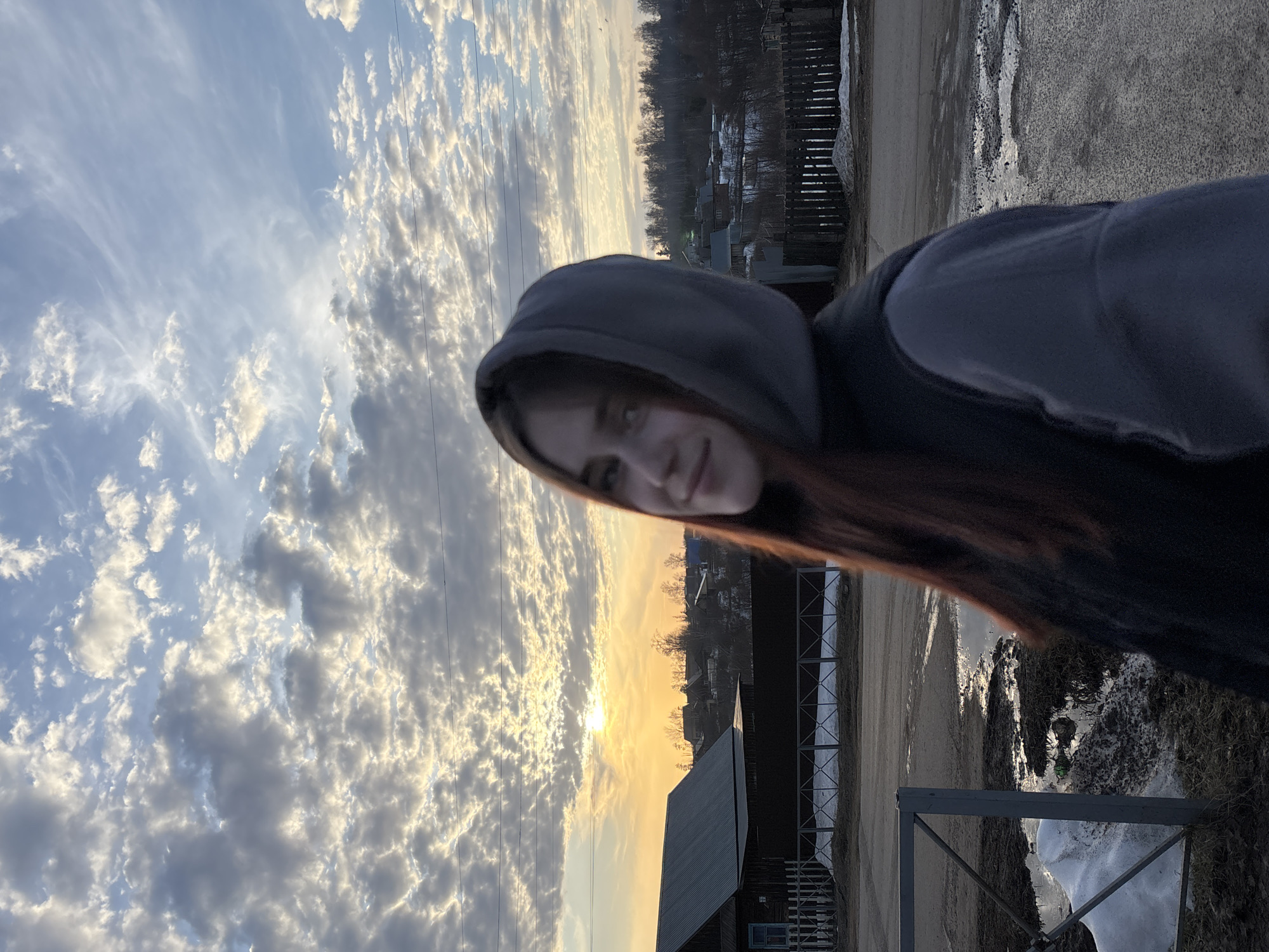 A waist-up shot shows a person with red hair, wearing a grey hoodie, positioned at an angle against a backdrop featuring a sunset and urban landscape. The sky is filled with clouds, and the sun is visible in the distance. The background includes buildings, trees, and a road, suggesting the location is within an urban area. There is snow on the ground, and there is also a metal fence. The location is the city of Фоки, Russia.