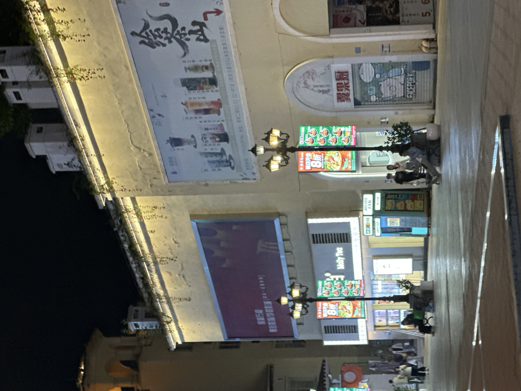This is a night-time, eye-level, medium shot of a pedestrian area in the Chongqing High-tech Zone, China. The scene is dominated by a long, light-colored building facade with several illuminated signs and advertisements.
On the left, a large, dark red billboard is mounted high on the building. Below it, a lamppost with three lights stands in front of a row of illuminated storefronts. Two vertical signs hang from the lamppost, one with a red background and Chinese characters indicating an "8% discount" on food, and another green sign with images of food. To the right of these signs is a storefront labeled "Molly Tea."
Further to the right, there is an arched entrance with a closed metal shutter featuring a cartoon drawing of a pig-like character and the text "FACEPORK." Next to this entrance, another storefront's details are obscured. A sign to the far right displays the numbers "5 6".
Several people are present in the scene. Three individuals are standing near the left edge of the image, their faces obscured. Two people are walking away from the camera on the left. One person is sitting on a bench in front of the "Molly Tea" store, and another individual is seated on the ground next to the lamppost. Two people are standing further to the right, near the entrance with the cartoon pig. The ground is paved and reflects the light from the buildings and signs. The overall ambiance suggests a commercial or entertainment district at night.