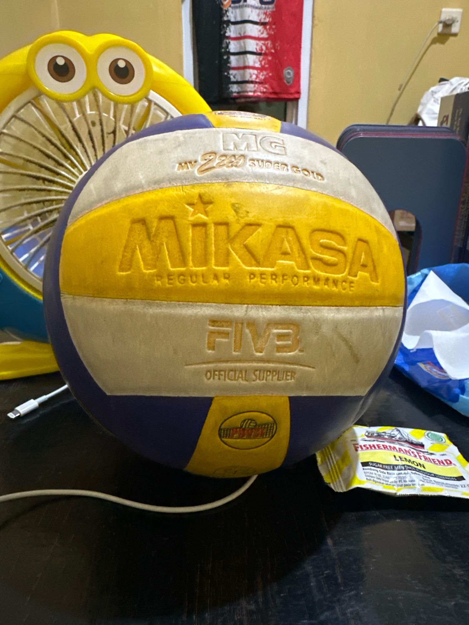The image displays a close-up, low-angle shot of a Mikasa volleyball placed on a dark, textured surface, likely a table. The volleyball is predominantly yellow and purple, with white sections bearing embossed text. Prominently visible are the "MIKASA" logo in large, yellow, embossed letters and "REGULAR PERFORMANCE" below it. Further down, "FIVB" is embossed in white, with "OFFICIAL SUPPLIER" underneath. A yellow fan with a cartoonish face and large brown eyes is partially visible behind the volleyball. To the right of the volleyball, a packet of "FISHERMAN'S FRIEND LEMON" lozenges is visible. A white charging cable with a silver connector is plugged into something off-camera and lies on the surface near the volleyball. The background is blurry and shows what appears to be a yellow wall and a red and white striped fabric hanging. There is no indication of a specific location within the city of Saruni, Indonesia.