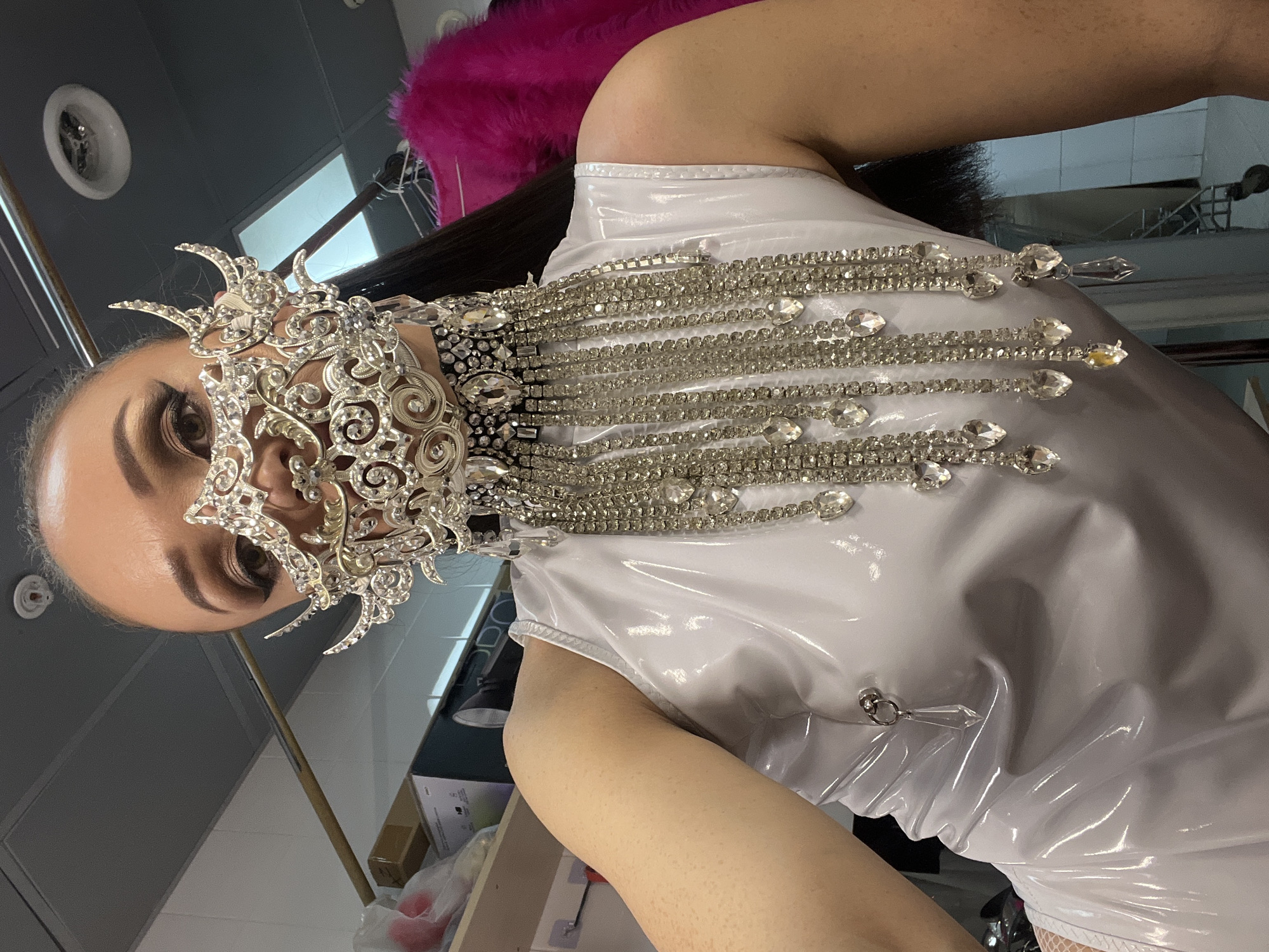 A person wearing a white dress with crystal embellishments and a silver mask is shown. The background suggests an interior space, possibly a dressing room, due to the presence of a clothing rack and a vent on the ceiling. The image appears to have been taken in Kazan, Russia, based on metadata not provided.