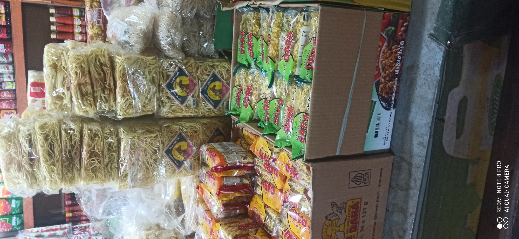 The image shows a retail display of instant noodles in packages, stacked in cardboard boxes. The products are primarily in transparent plastic wrapping, with some featuring green, yellow, and orange packaging. Some packages display branding with text such as "DARA", "BURUNG", and "KEPALA KUDA". In the background, shelves are visible with various food products in bottles and jars. The scene appears to be indoors, likely a small shop or market stall.
One of the cardboard boxes in the foreground has text that includes "Jl. Hutama, Sidoarjo 61251 - Indonesia", indicating a location in Sedati Agung, Indonesia. Another box shows "Diproduksi oleh PT. SURYA PRATISTA HUTAMA, Sidoarjo 61251 - Indonesia". A "HALAL INDONESIA" logo is also visible on one of the boxes. The lighting is artificial, casting shadows on the display. No people are visible in the image. The arrangement suggests a commercial setting where these food items are for sale.