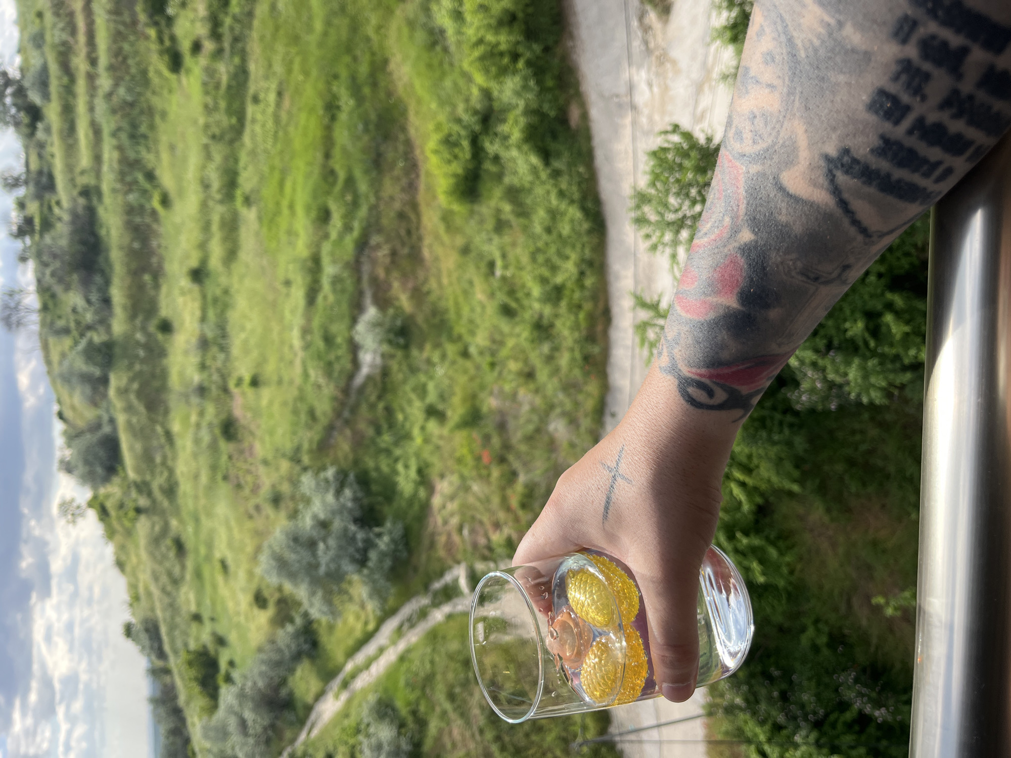 The image depicts a person's arm with a tattoo holding a glass filled with yellow and brown objects, possibly candies or chocolates. The person is standing on a balcony with a metal railing. In the background, there is a grassy hillside or a park with a pathway. The location appears to be elevated, offering a view of the landscape. Based on visual cues, the image could be taken somewhere in the city of Varna, Bulgaria.