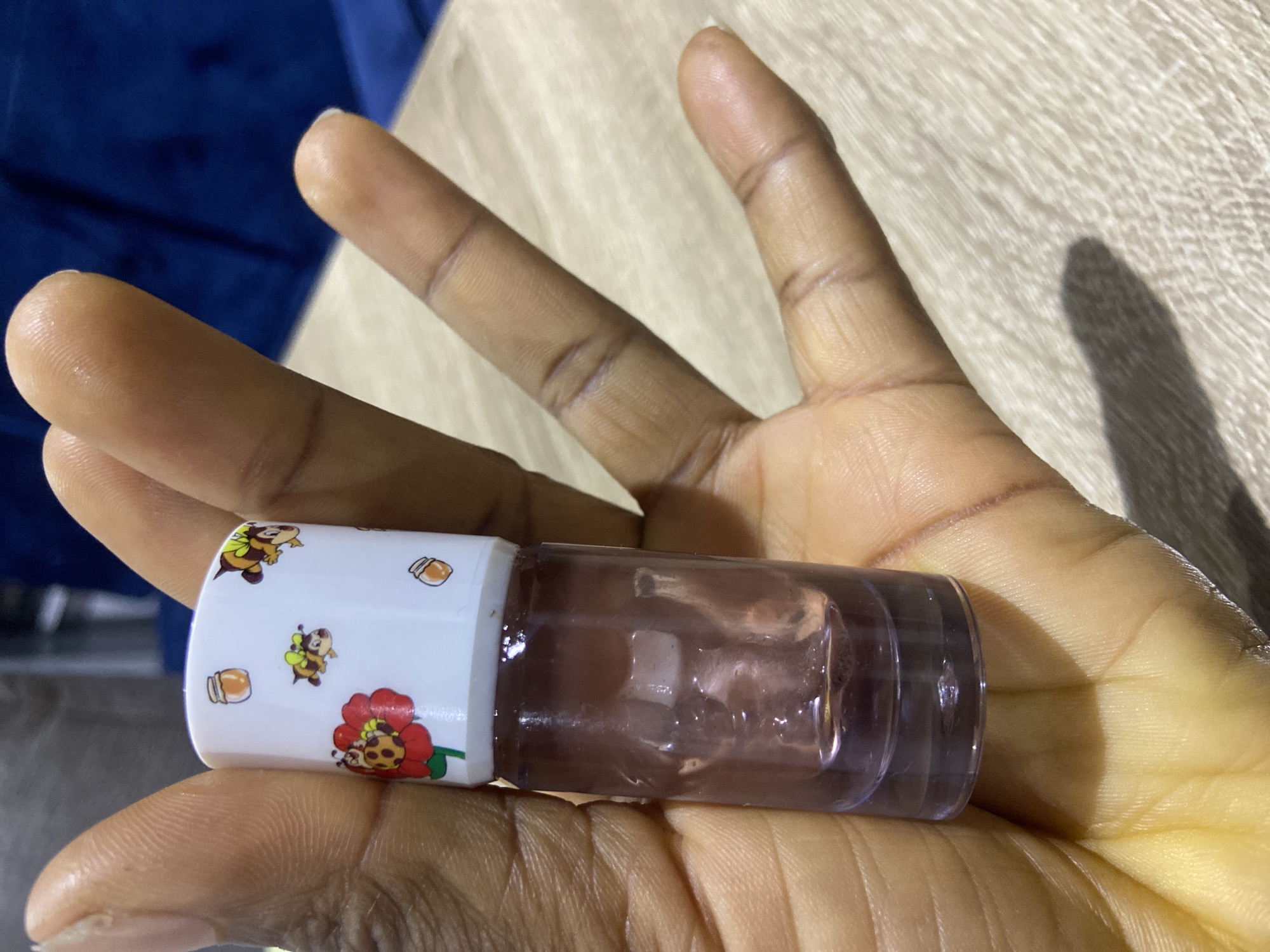 An overhead, close-up shot shows a hand in Gura Top, Nigeria, holding a small, clear glass bottle filled with purple liquid. The bottle has a white cap adorned with cartoon bees, a ladybug on a red flower, and small jars of honey. The hand, with visible lines and creases, supports the bottle from below, with fingers spread. The background features a light-colored wooden surface and a dark blue blurred object, possibly furniture.
