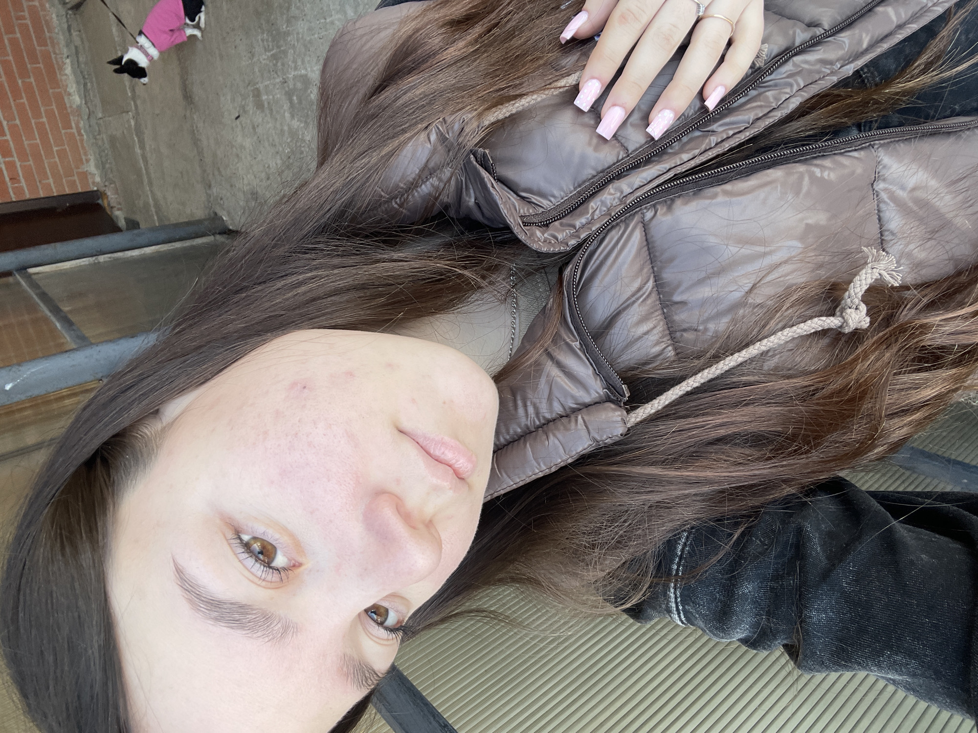 The image features a close-up, angled selfie of a young female taken outdoors in Фоки, Russia. She has long, dark brown hair, light brown eyes, and visible skin imperfections on her face. She is wearing a dark brown, glossy puffer vest over a dark garment, possibly a black denim jacket, with a silver chain necklace visible beneath. Her left hand, positioned on her chest, displays long, square-tipped light pink fingernails with white embellishments on some nails, and two rings (one silver, one gold) on her ring finger. In the upper right background, a small black and white dog wearing a pink coat and pink collar is visible on a leash, standing on a concrete surface next to a red brick wall and a dark door. A ribbed, light-colored panel or wall is present in the lower left background.