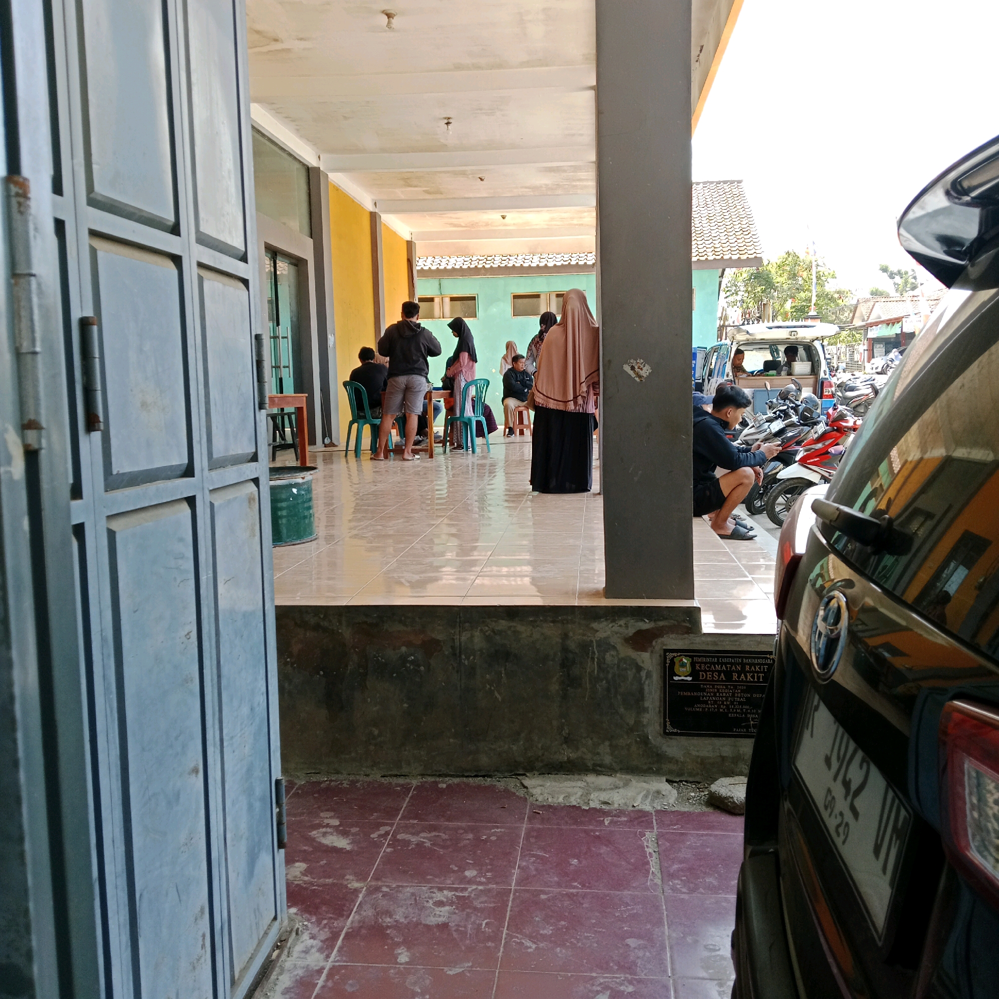 The image depicts an outdoor scene in front of a building with a yellow and green facade, possibly in the city of Rakit, Indonesia, as suggested by a sign in the lower left corner that reads "PEMERINTAH KABUPATEN BANJARNEGARA KECAMATAN RAKIT DESA RAKIT". In the foreground, a dark-colored car is parked, partially obscuring the view of the building and its surroundings. The car has a license plate that reads "1942 UN 09-29".
The building appears to be a covered walkway or veranda, with a tiled floor. Several people are gathered in this area, some seated on green plastic chairs around a table, and others standing. One individual is wearing a black hoodie and grey shorts, another is in a pink outfit with a hijab, and a third person wears a brown hijab and a black dress. Another group is visible further back, near a van parked outside. A stack of motorcycles is parked to the right of the van. The overall atmosphere suggests a casual gathering or waiting area. The environment is bright, indicating daylight.