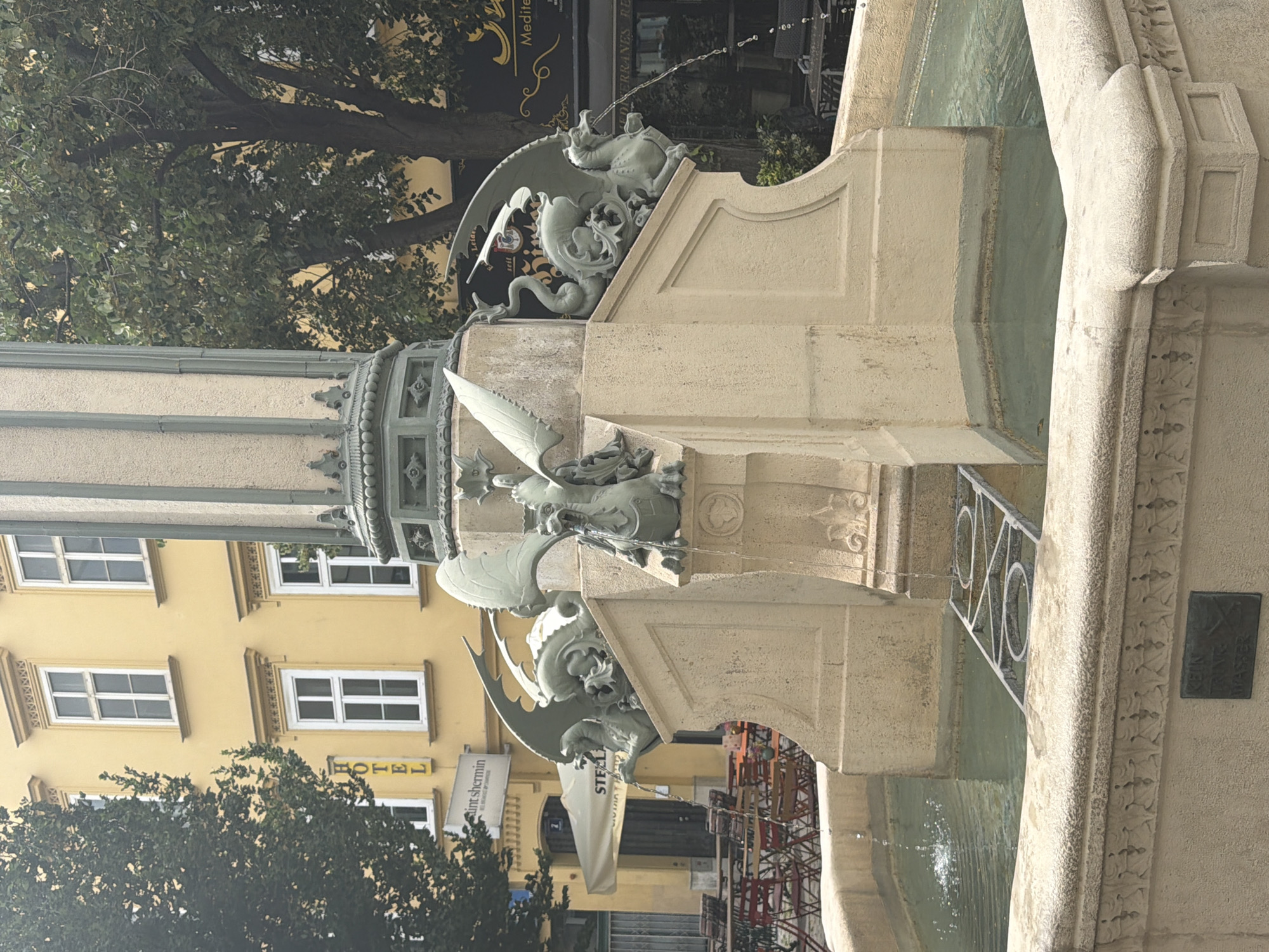 This media file depicts an outdoor scene in Vienna, Austria, featuring a stone fountain adorned with intricate dragon sculptures. The fountain is a multi-tiered structure with water cascading into a pool. One of the dragon sculptures, with outstretched wings and a serpentine body, is a prominent element. Behind the fountain, a building with a yellow facade and white-framed windows is visible. Trees with lush green foliage are also present in the foreground and background, suggesting a daytime setting, possibly in a park or public square.
A sign with the word "HOTEL" in yellow lettering is affixed to the yellow building. Another sign, partially visible, reads "saint shermin" and "STEEL". The architectural style of the building and the elaborate design of the fountain suggest a historical or cultural location within the city. The presence of construction scaffolding in the background indicates ongoing development or maintenance in the vicinity. The scene appears calm and serene, with no people readily visible in the frame. The water in the fountain is clear, and the lighting suggests it is daytime, with the sun casting shadows that are not intensely harsh. The overall impression is of a picturesque urban landscape with elements of art and architecture.