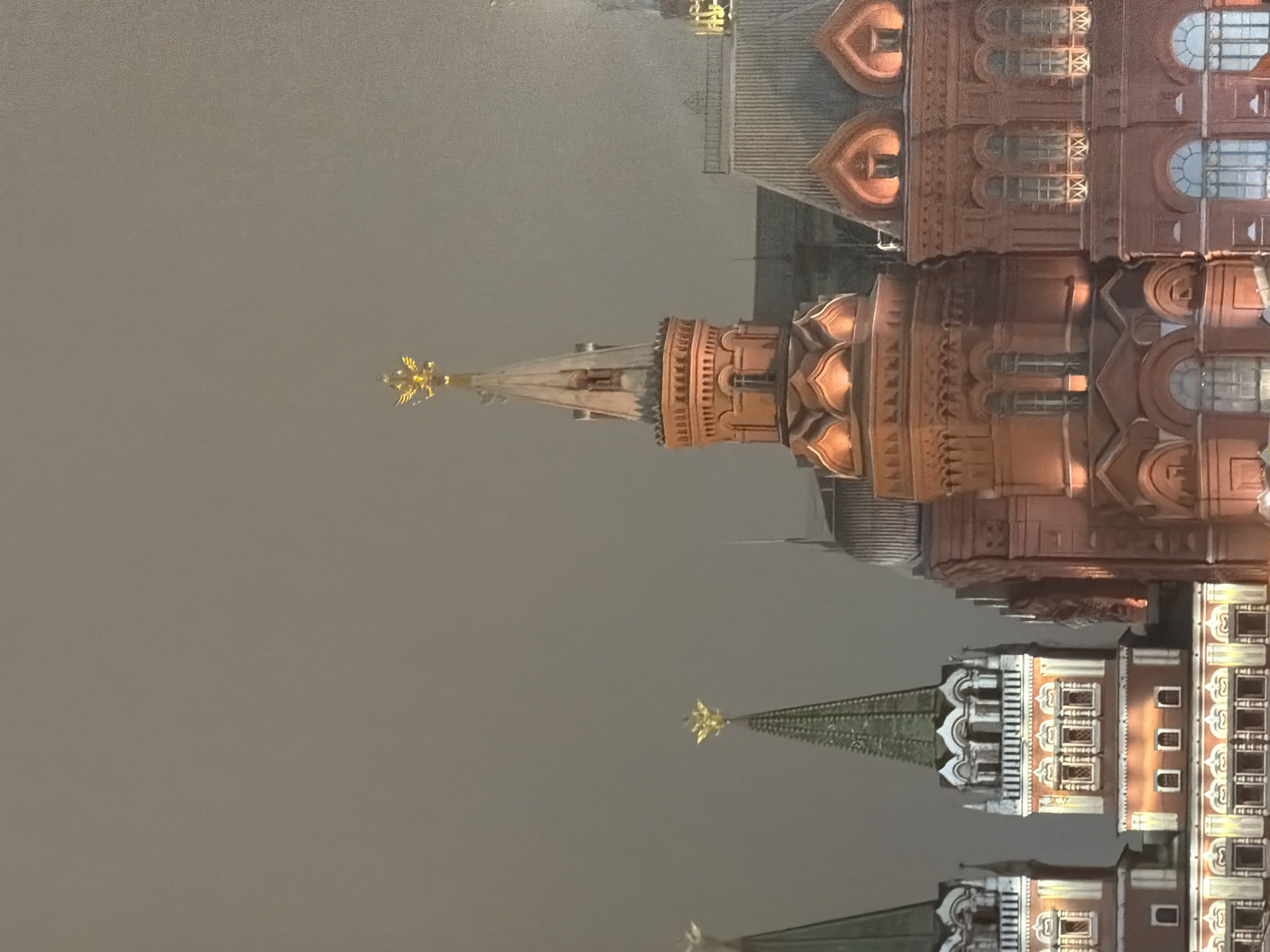The image depicts a portion of the Moscow Kremlin at night, featuring several towers with distinctive architectural details. The towers are illuminated, showcasing their intricate designs and ornamentation. The surrounding environment appears to be shrouded in fog, obscuring the details of the background. The towers are adorned with golden stars atop their spires.