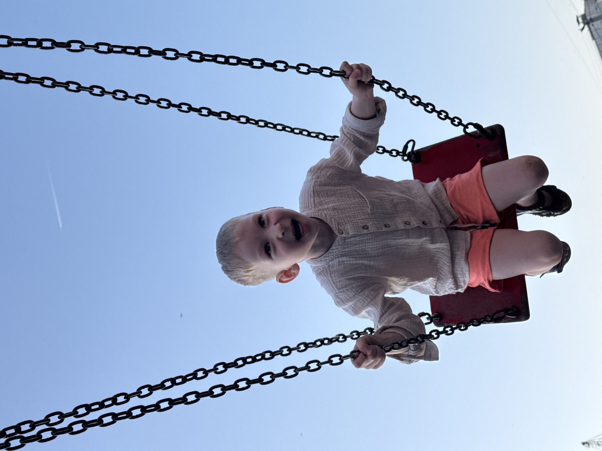 A low-angle shot depicts a child mid-swing, suspended against a clear, light-blue sky. The child is wearing a light-colored, long-sleeved button-up shirt and orange shorts, along with black shoes. The swing seat is red, and the swing is held up by heavy, black chains. The child is gripping the chains with both hands and appears to be looking down, with a wide, excited expression. There is a faint contrail from an airplane visible in the upper left of the frame. The location details, Chaikovsky, Russia, cannot be verified through the image alone.