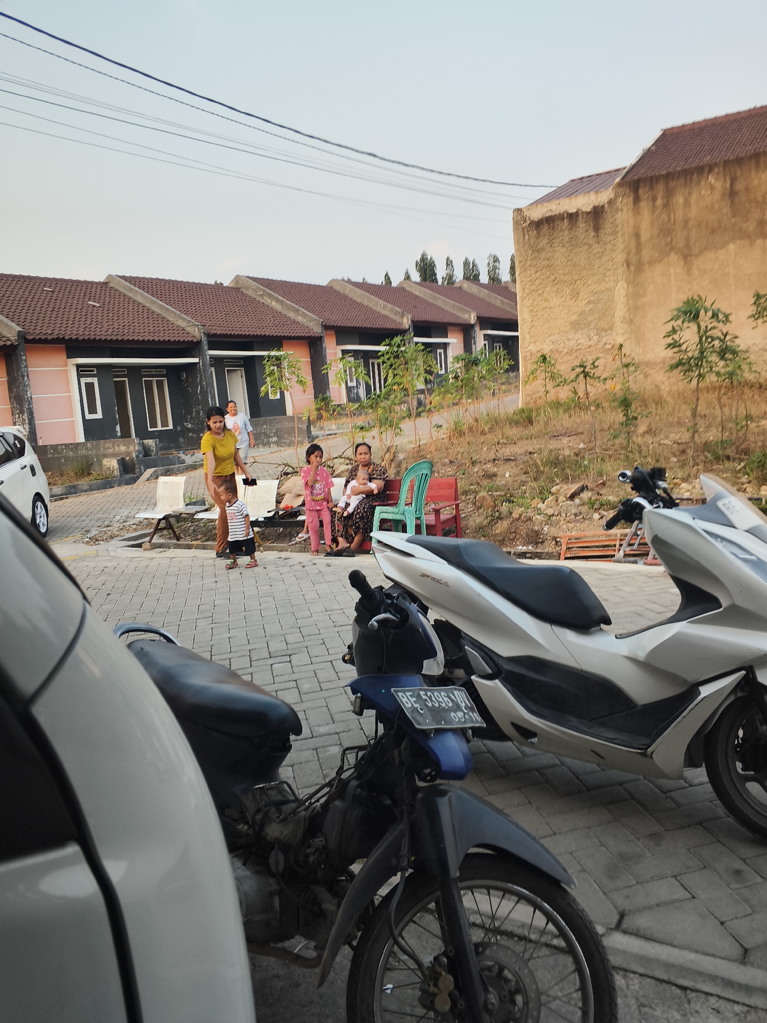 In the city of Banjar Agung, Indonesia, this image captures a residential street scene. The foreground is dominated by the front of a white car and two motorcycles parked on a cobblestone pavement. The motorcycle on the right is white, and the one on the left is dark blue with a visible license plate. In the background, several houses with brown tiled roofs are visible, characterized by their uniform design and simple facades with dark windows and doors. A light brown, textured wall of a building is prominent to the right. Several individuals are present in front of one of the houses. A woman in a yellow shirt stands near a young boy in a striped shirt. Another woman is seated, holding a baby, next to a young girl wearing a pink outfit. A third woman stands behind the seated woman. The environment suggests a residential area with a clear sky and some sparse vegetation.