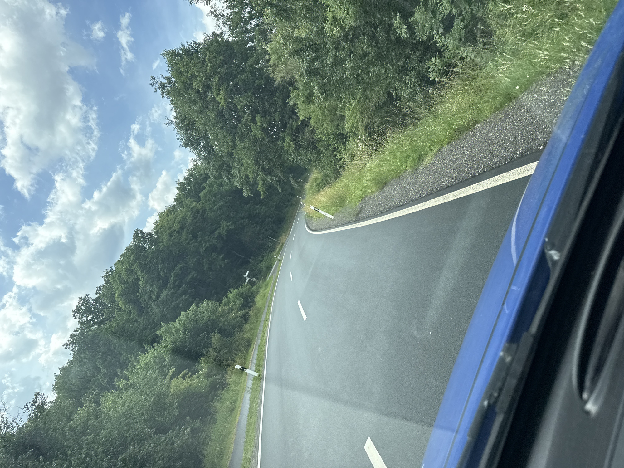A car-mounted, eye-level, medium shot captures a view of a paved, winding road cutting through a dense forest. The road, with white lane markings, curves to the right, disappearing into the greenery. The forest, comprised of various shades of green trees, lines both sides of the road, with a strip of tall grass and gravel bordering the right side of the asphalt. The sky above is a bright blue, dotted with fluffy white clouds. A small section of the blue car's hood is visible in the foreground. The scene suggests a drive through a rural, wooded area.