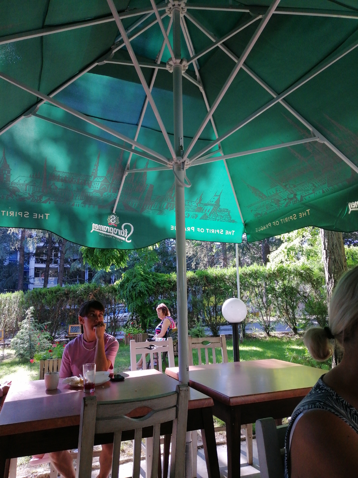 A group of people are dining outdoors under a large green umbrella. The umbrella is decorated with an illustration of Prague's skyline and the text "THE SPIRIT OF PRAGUE". In the foreground, a woman with dark hair sits at a table, looking thoughtfully to the side with her hand on her chin. A partially consumed meal and drink are on the table in front of her. Further back, a woman with blonde hair in a ponytail is seated at another table, facing away from the camera. The dining area is set within a lush, green garden, with trees and manicured hedges visible in the background. The overall atmosphere suggests a relaxed outdoor dining experience.