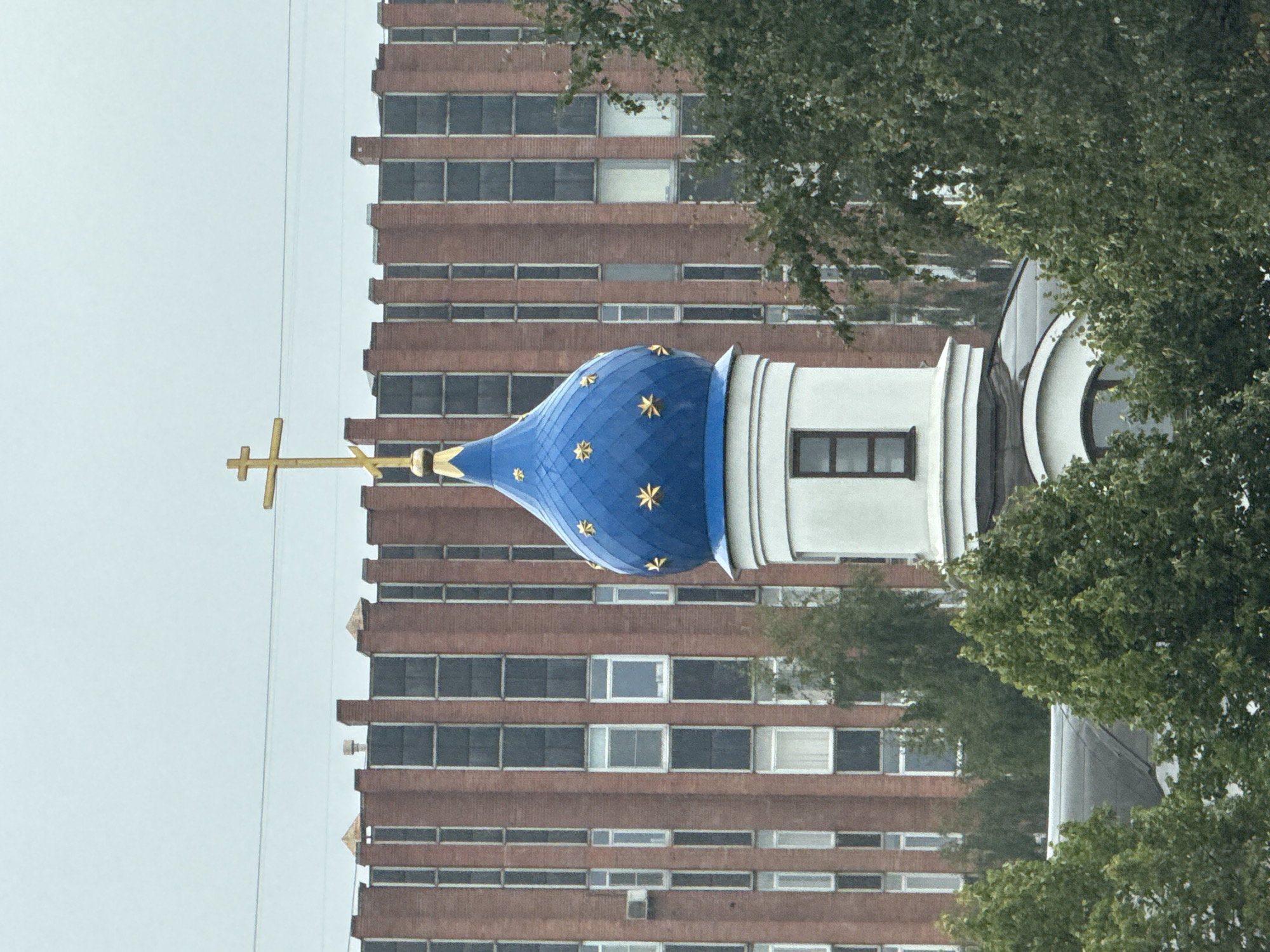 The image shows the dome of an Orthodox church with a golden cross against a backdrop of a red brick apartment building under a cloudy sky. The dome is painted bright blue and adorned with golden stars. A white cylindrical section below the dome has a small rectangular window. In the foreground, lush green trees partially obscure the lower portion of the church structure. The apartment building behind the church features numerous tall, narrow windows arranged in vertical columns. The overall impression is one of a religious structure situated within a modern urban environment. Without specific identifiable landmarks, pinpointing the exact location in Saint Petersburg is not possible based solely on the visual information provided.