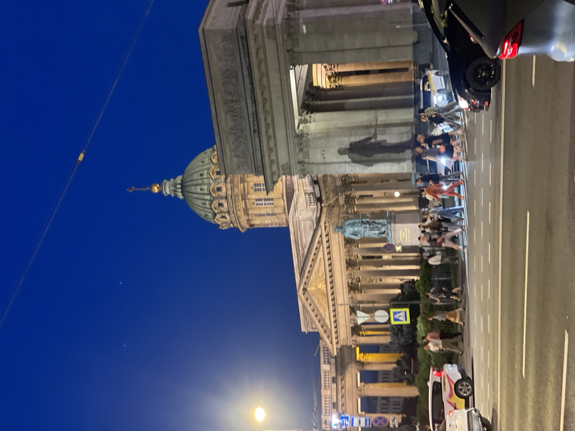 The image captures a nighttime urban scene in Saint Petersburg, Russia, featuring the Kazan Cathedral under a deep blue twilight sky. Overhead wires are visible across the upper portion of the frame. The Kazan Cathedral is prominent in the midground, distinguished by its large green dome topped with a golden cross, and a grand portico supported by columns. In front of the cathedral stands a bronze statue of Mikhail Kutuzov. To the right of the cathedral, a neoclassical arched structure with columns is visible.
In the foreground, a multi-lane street features white pedestrian crosswalk markings. Approximately 15-20 pedestrians are crossing the street in both directions on the marked crosswalk, illuminated by streetlights. Traffic includes a partially visible white SUV with yellow and red markings on the left, a dark sedan stopped at the crosswalk on the right, and a partially visible gray car's rear section. Another white car is visible further down the street behind the pedestrians. Various road signs, including a pedestrian crossing sign, are visible near a landscaped median.