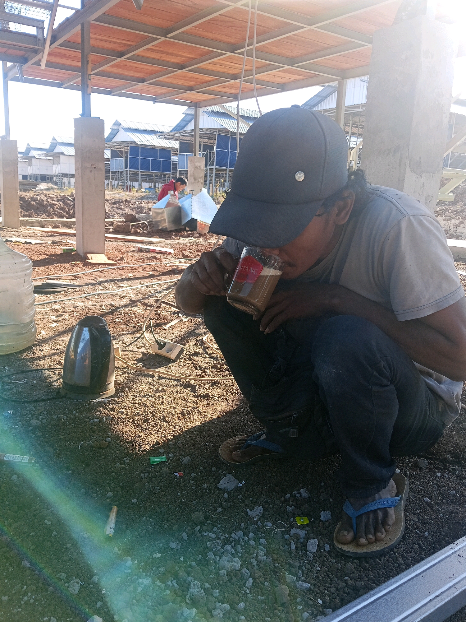 The image depicts a construction site in Kecamatan Lewa, Indonesia, under a bright, sunny sky. In the foreground, a person wearing a grey t-shirt, dark pants, a dark baseball cap, and blue flip-flops is squatting and drinking from a clear glass. The glass contains a light brown beverage with foam on top, and a red label with white lettering is visible on its side. To the left of the person, a large clear water jug and a black electric kettle are placed on the ground, with electrical cords and a power outlet visible. The ground is covered in dirt and gravel. In the background, several unfinished structures with metal frames and wooden ceilings are present, along with rows of buildings with blue tarps and silver roofs. Another person in a red shirt is visible further in the background, near a pile of dirt. Debris, including a crumpled cigarette pack and a green wrapper, are scattered on the ground. The overall scene suggests a break or pause during construction work.