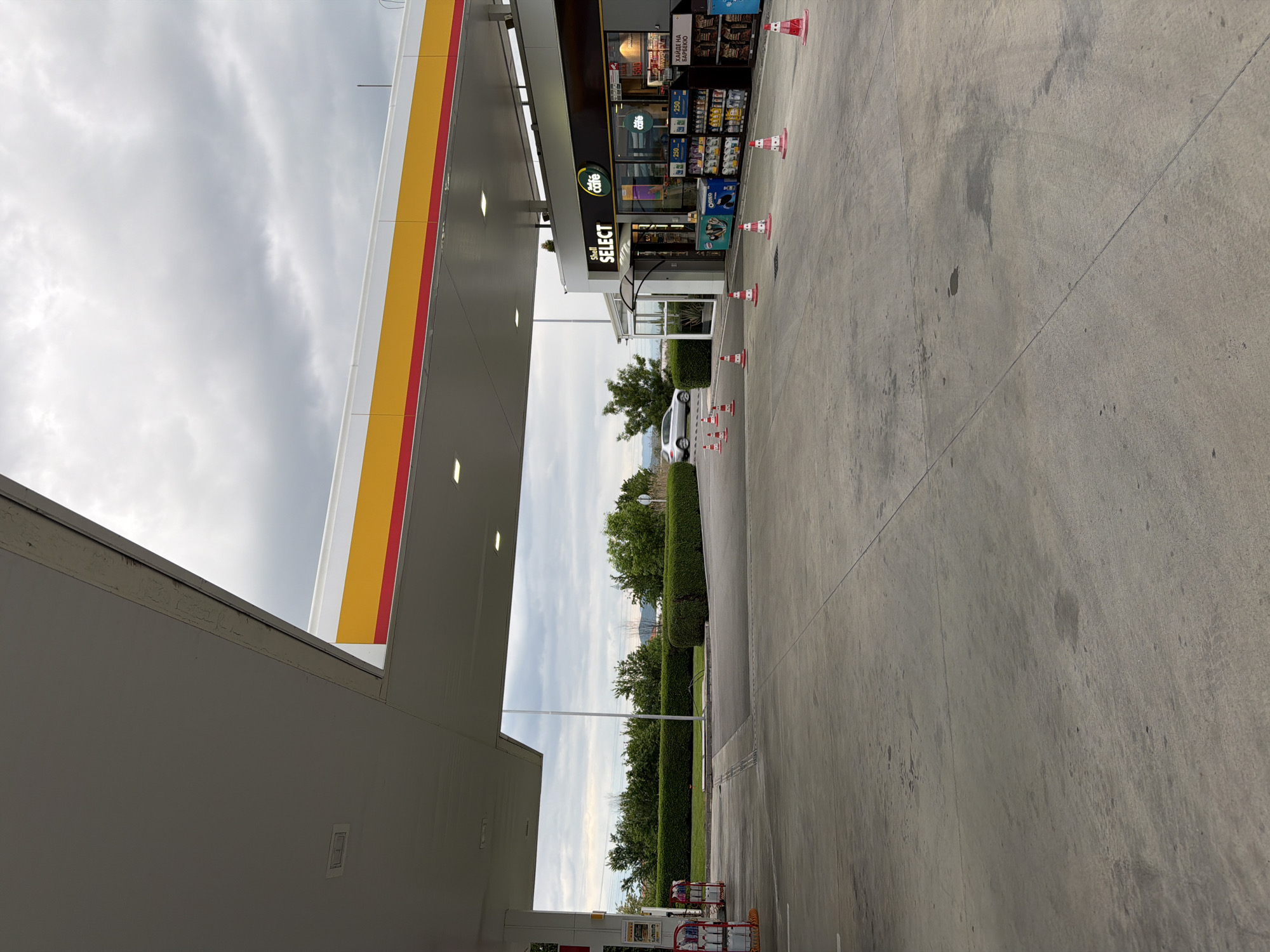 The image depicts a Shell gas station in Dragor, Bulgaria. The sky is overcast. A Shell Select convenience store is visible with an assortment of snacks and beverages. A white car is parked beyond the store. Orange and white cones are placed near the store entrance. Green hedges are present in the background. The concrete ground appears to be stained.