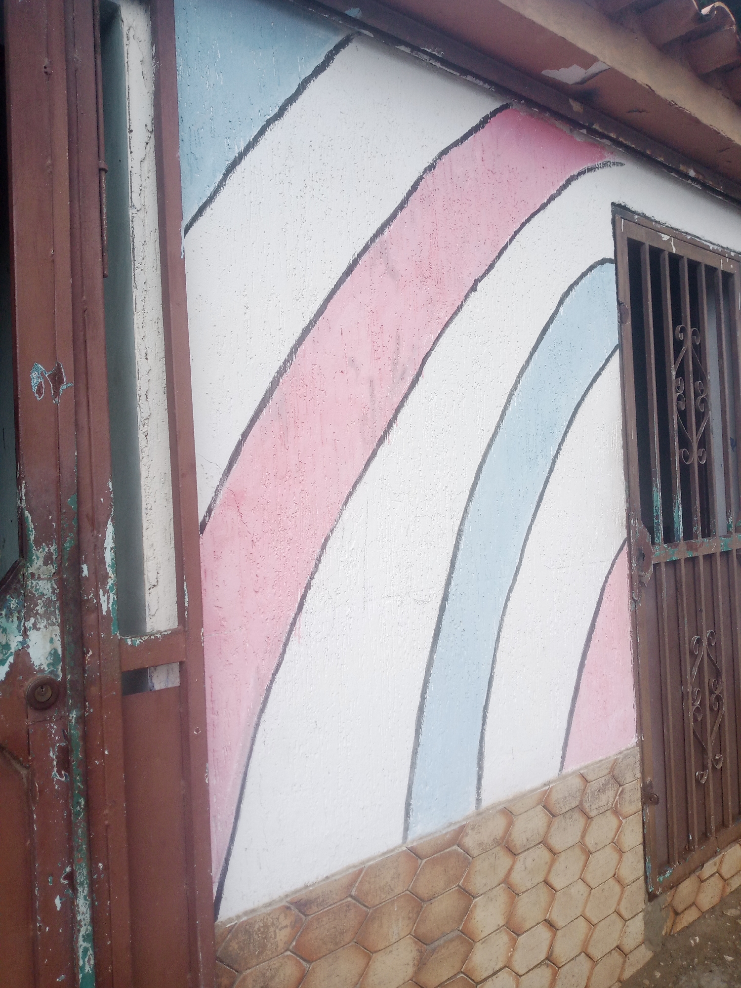 This is a medium-angle, eye-level shot of the exterior wall of a building in Santa Ana, Venezuela. The wall is painted with a vibrant, abstract design of curved stripes in white, pink, and light blue, outlined in black. The paint is textured, giving the stripes a slightly raised appearance. Towards the bottom of the wall, there's a band of hexagonal tiles in shades of brown and tan.
To the left of the painted wall is a weathered, reddish-brown metal door or gate. It appears to be partially open, revealing a glimpse of something behind it. The metal shows signs of rust and peeling paint, with some greenish-blue patches visible.
To the right of the painted wall is a window with a dark, ornate metal grate. The grate is also a reddish-brown color and shows some signs of age and weathering. The wall directly above the window is partially visible, covered by a brown tiled roof.
The overall impression is of a building with some signs of age and wear, but with a cheerful and colorful artistic touch on its facade. The lighting suggests it could be daytime, possibly overcast given the diffused light and lack of harsh shadows. There is no visible text in the image.