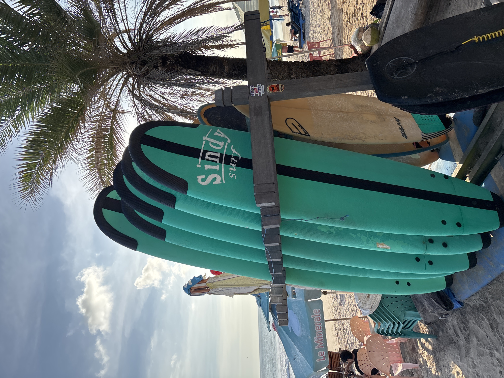 The image depicts a beach scene in Kuta, Indonesia, featuring a stack of green surfboards labeled "Sindy Surf" resting against a wooden rack. Adjacent to the surfboards, there is a tan-colored surfboard and a black bodyboard. Palm trees cast shadows across the scene, and in the background, the beach is visible with beach chairs and people. An advertisement for "Le Minerale" is seen as well.