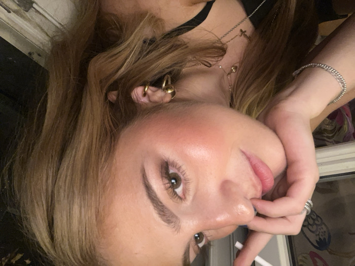 The image is a close-up, high-angle portrait of a young female. The subject has light brown hair, brown eyes, and is wearing makeup, including blush and mascara. Her right hand is positioned along her jawline, with fingers curled, and a white cylindrical object, consistent with a cigarette or vape pen, is held between her right thumb and index finger, angled upwards near her mouth. She wears multiple silver-colored rings on her right ring finger and a silver-colored beaded bracelet on her right wrist. Her visible left ear features at least three gold hoop earrings. She wears two layered gold-colored necklaces: a shorter one with a small charm and a longer one with a small cross pendant. Her attire includes a dark, thin-strapped top. The background on the left shows a window frame with a light-colored sill, behind which a blurred, patterned interior element is visible. On the right, a dark vertical surface with a lighter trim is present. The indoor lighting appears warm. The specific location within Paris, France, cannot be determined from the visible background elements.