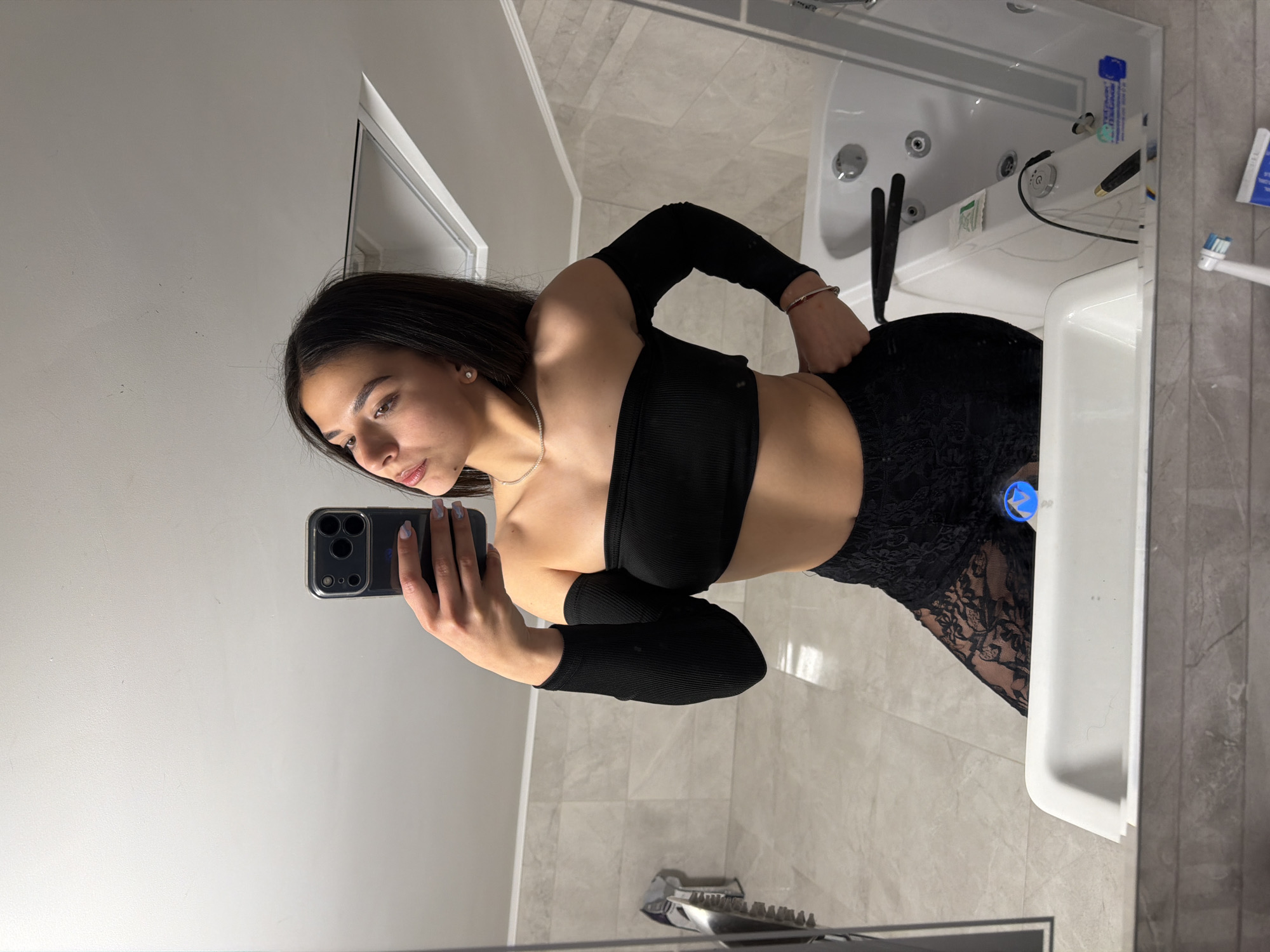 A woman is taking a selfie in a bathroom. She is wearing a black crop top and black lace pants. She is holding a black smartphone in her left hand. The bathroom has gray tile walls and a white sink. There is a bathtub with jets in the reflection of the mirror. The location of the image is not specified and there is no information about it to confirm it. The city of Gabrovo, Bulgaria, is also not mentioned.