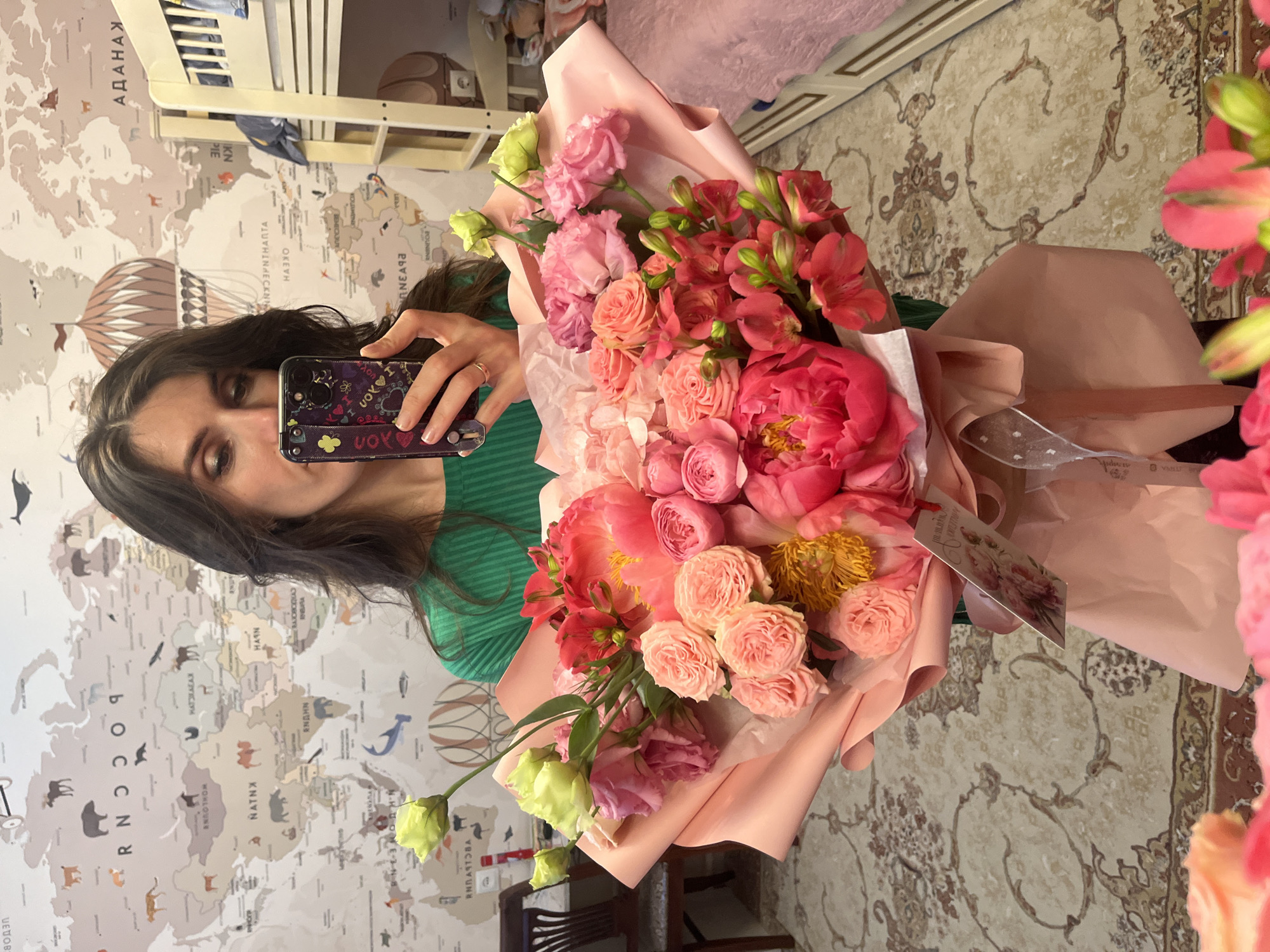 Here is a detailed description of the image:
**People:** A woman with long, dark hair and a green striped shirt is visible in the image. She is holding a smartphone up to take a picture of herself in a mirror. She wears a ring.
**Subjects:** The main subject of the image is a large bouquet of pink and peach-colored flowers. It includes roses, peonies, and alstroemeria.
**Objects:** Key objects include the bouquet of flowers, the woman's smartphone with a purple case, a bed with a ladder, a chair, and a mirror. The smartphone case has a decorative strap with writing on it. There is a gift tag on the bouquet, and a floor with a patterned carpet. There is a world map on the wall behind the woman.
**Actions:** The woman is taking a selfie with the bouquet of flowers.
**Interactions:** The woman is interacting with her smartphone to capture an image of herself and the flowers.
**Environment:** The location appears to be a room within a home or apartment. The world map is likely a decorative element on the wall.
**Location:** The location is likely Kaspiysk, Russia based on the writing on the wall map.