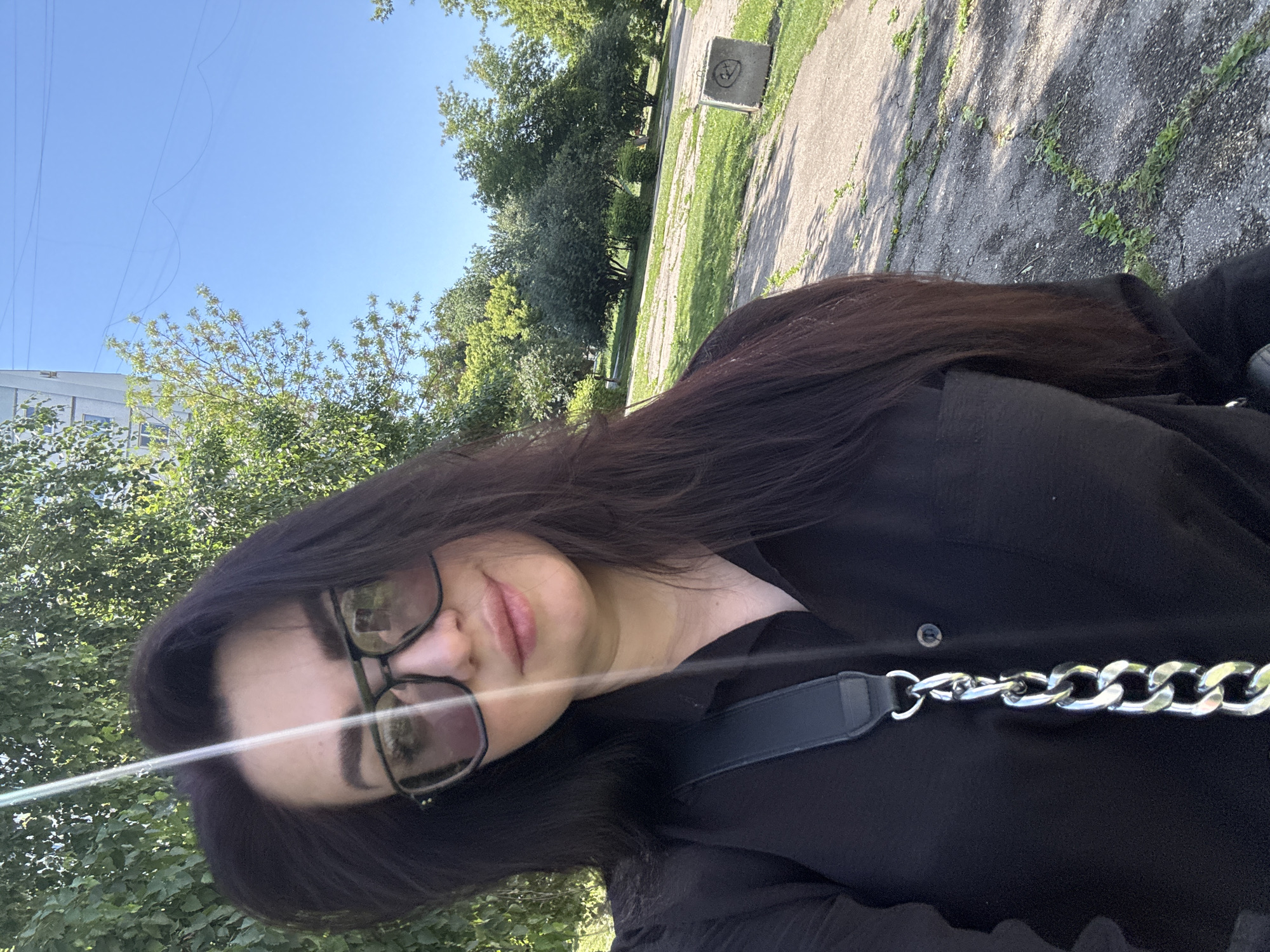 The media file is a close-up, self-portrait photograph taken outdoors during the day. The subject is a person with long, dark hair, wearing glasses and a black top. They are looking towards the camera, with their face partially obscured by their hair. A thick, silver chain is visible across their chest, attached to a black strap.
The background of the image shows a blurred scene of trees and a concrete path, suggesting an outdoor setting such as a park or a walkway. The lighting indicates it is daytime, likely sunny, given the brightness and shadows. The overall mood appears casual and relaxed. There is no visible text in the image.