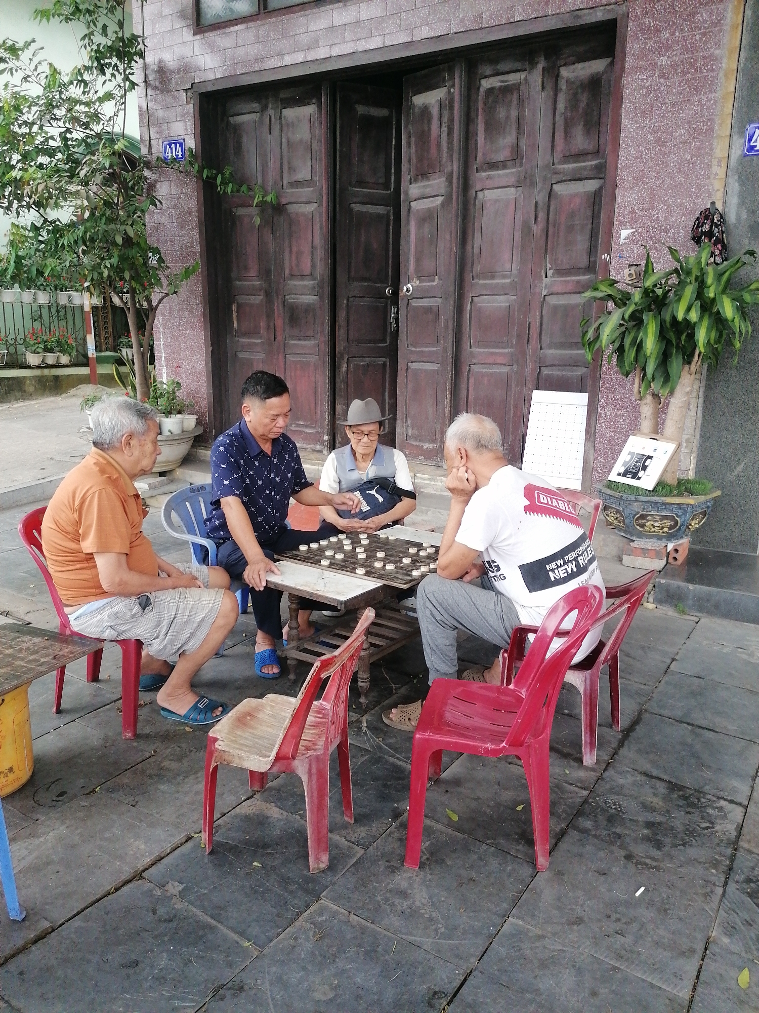 Four elderly men are engrossed in a game of checkers or a similar strategy game. They are seated on an outdoor patio or sidewalk in front of a building with a large, dark wooden door. The location is identified as Ha Tu Ward, Vietnam. The men are seated on red plastic chairs around a low table with the game board. Various plants and greenery are around them.