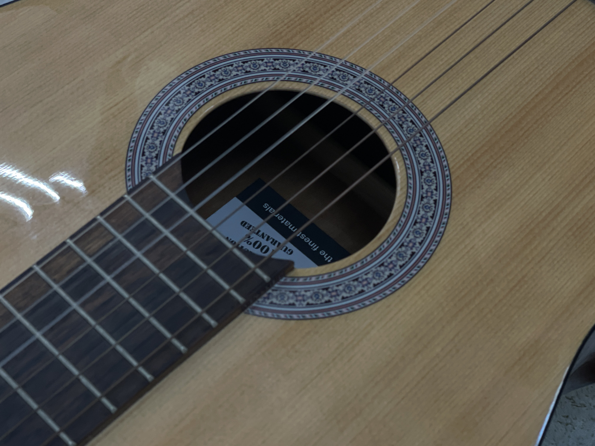 The image shows a close-up of a classical guitar, with the focus on the soundhole and part of the fretboard. A label is visible inside the soundhole with the text "100% GUARANTEED the finest materials". The guitar has a light wood finish and decorative inlay around the soundhole. The lighting suggests an indoor setting. There is no information available to place this image within the city of Ulmen, Germany.