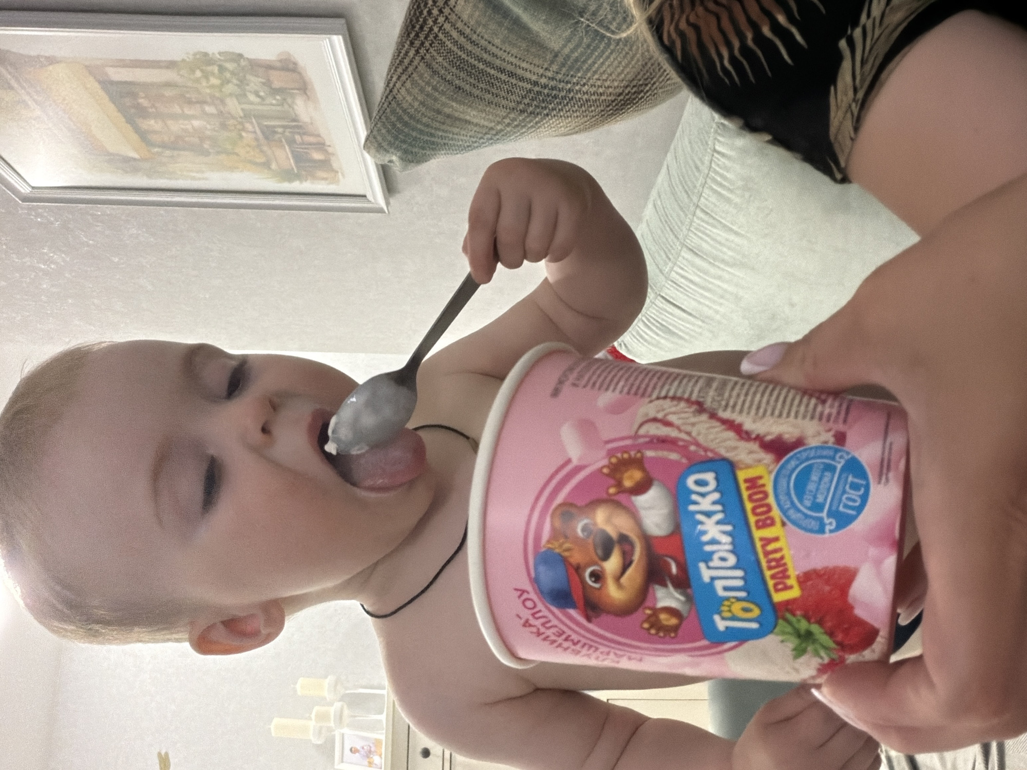 A baby is eating yogurt with a spoon. The yogurt container is pink and features a cartoon bear. The image was taken indoors. In the background, there is a framed picture and a couch. There is no information in the image to suggest it was taken in the city of Votkinsk, Russia.
