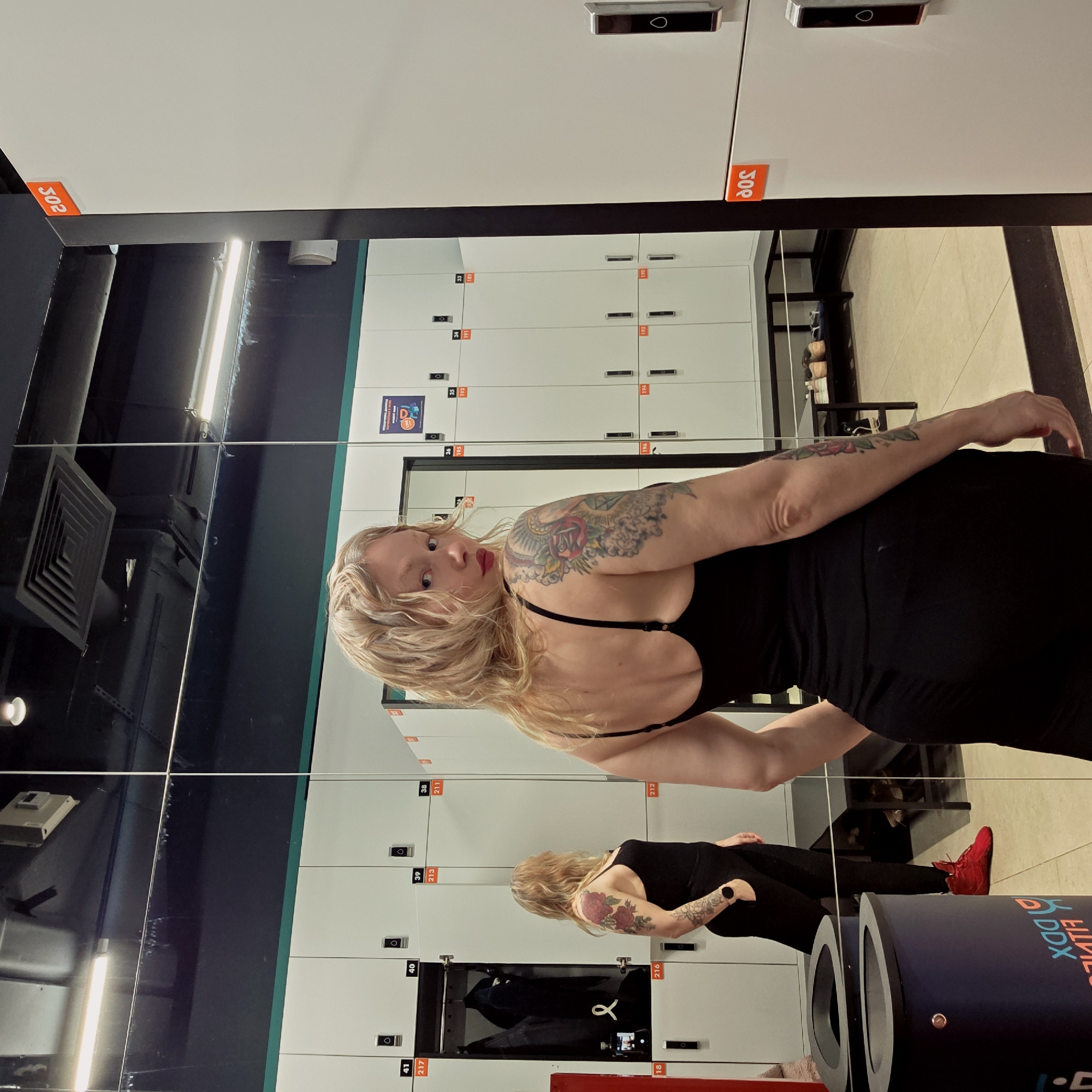 A photo taken at a fitness center in Saint Petersburg, Russia, shows a woman reflected in a mirror wall. The woman has blonde hair, fair skin, and is wearing a black dress. A visible tattoo decorates her upper arm. Behind her, lockers line the wall. The lockers are white with orange number tags, which are between the numbers 302 and 418. Some lockers have doors open, revealing dark items inside. The environment is brightly lit with fluorescent lights, and part of the gym's branding, including a blue and white logo, is also visible at the bottom of the frame.