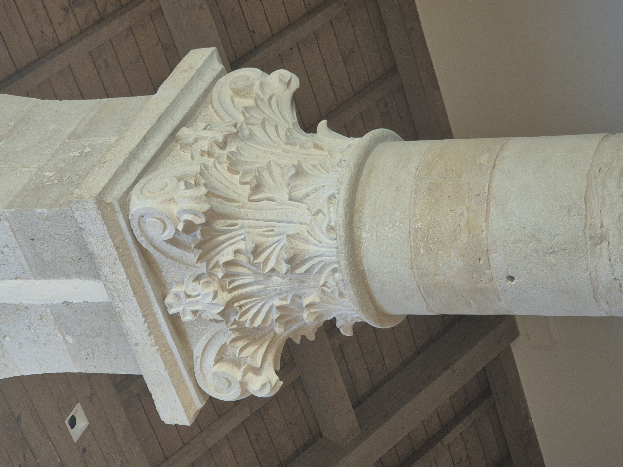 The image shows a close-up of a stone pillar with ornate carving on its capital. The pillar is part of the architecture of the Crusader Monastery of Abu Gosh, located in Abu Gosh, Israel. The capital is decorated with acanthus leaves and volutes, a style characteristic of Romanesque architecture. Above the pillar, wooden beams and ceiling planks are visible, along with a square light fixture embedded in the ceiling. The pillar itself is made of roughly dressed stone blocks. The overall scene suggests an interior or covered area within the historic monastery.