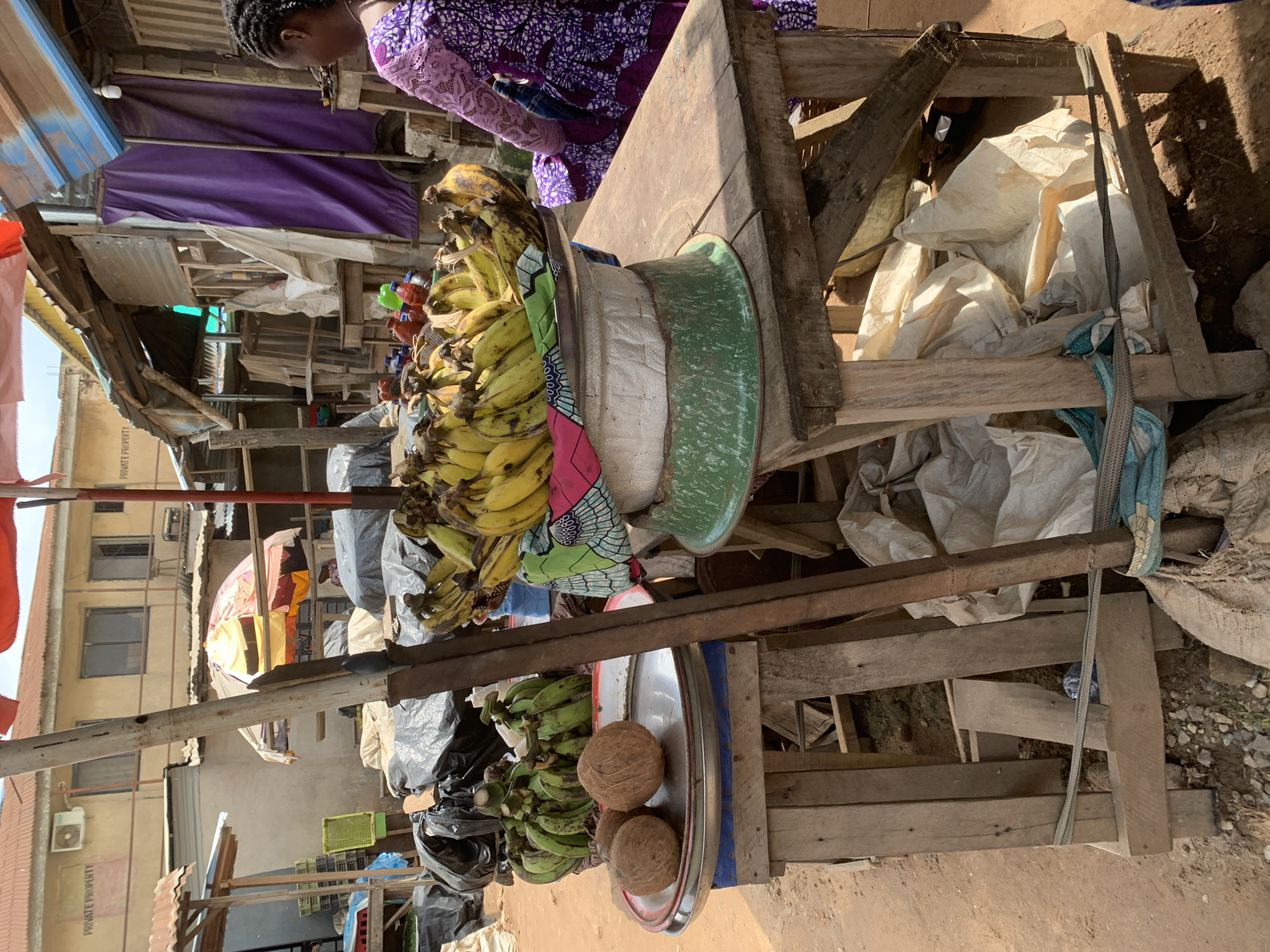 The image shows a market stall in Shagamu, Nigeria, from a slightly elevated perspective. A woman in a purple patterned dress is visible in the upper left corner, partially obscured by the stall's merchandise. The stall is constructed of rough-hewn wood and contains a variety of fruits and vegetables, including bananas (some yellow, some green) and coconuts. These are displayed on metal trays and in a green enamel basin. Underneath the stall are large white sacks and other debris. The background shows a bustling market scene with other stalls, buildings, and a mix of residential and commercial structures. The lighting suggests it is daytime.