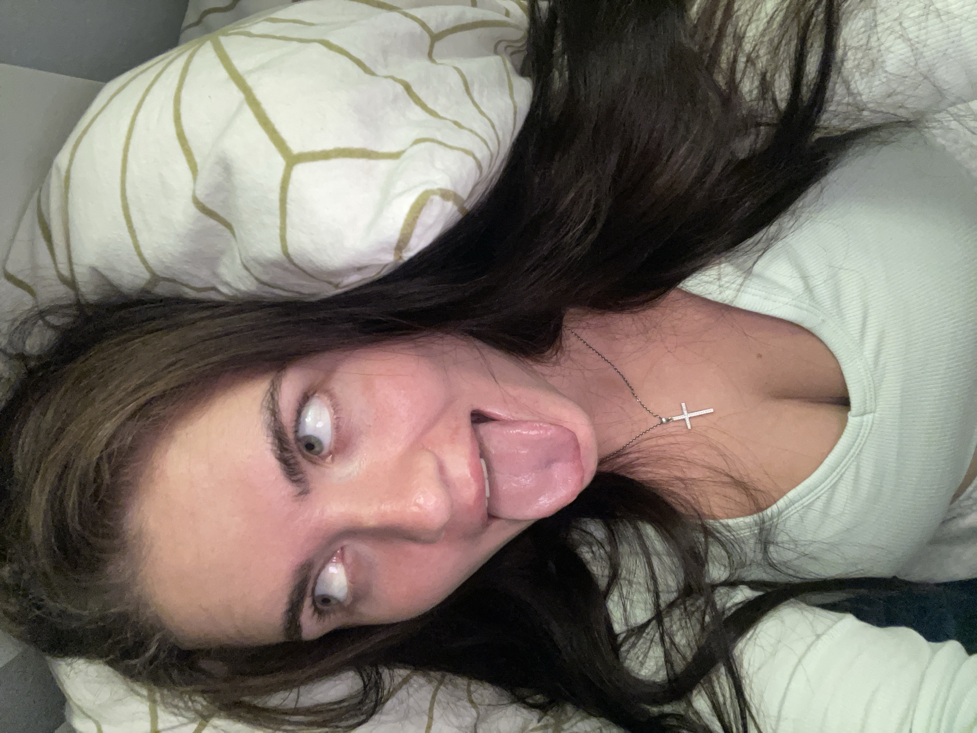 A young woman with long, dark hair is lying down, looking up and to her left with her tongue sticking out. She is wearing a light green, ribbed top with a cross necklace. She is on a pillow with a green and white geometric pattern. The background is out of focus, but appears to be indoors. The image was taken in Мосметаллоконструкция Онт, Russia.
