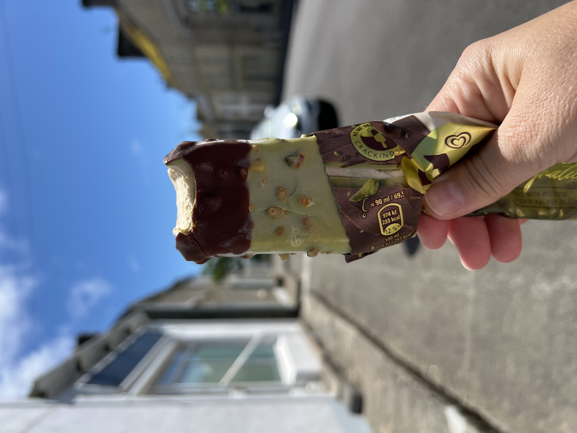 A hand holds a partially eaten pistachio ice cream bar covered in chocolate and chopped pistachios. The ice cream bar is in the foreground, with its wrapper partially peeled back, revealing nutritional information. In the background, a blurred street scene unfolds under a bright blue sky with scattered clouds. Buildings line the street on either side, and a white car is visible in the middle distance. The scene is indicative of a sunny day in Mountcharles, Ireland, likely on a residential street.