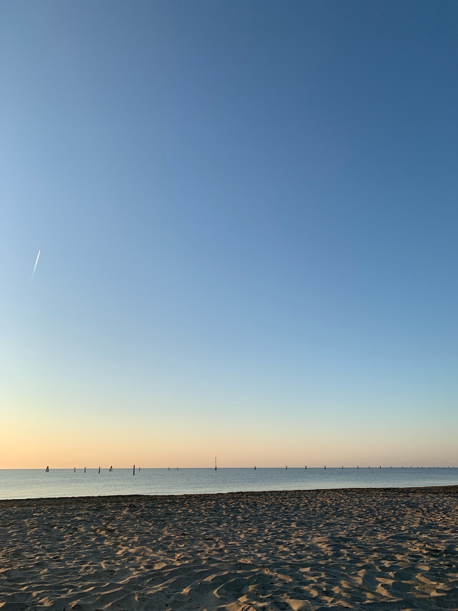 The image depicts a tranquil beach scene with a vast, clear blue sky dominating the upper portion. A faint contrail from an airplane is visible in the upper left quadrant. The horizon line separates the sky from the calm, light-colored sea, which stretches across the frame. A series of dark, slender poles or markers are scattered across the water, spaced at intervals. The foreground is composed of textured sand, showing ripples and indentations from the natural contours of the beach. The lighting suggests either early morning or late afternoon, with a subtle gradient of color in the sky near the horizon transitioning from pale yellow to blue. The scene is devoid of any human presence or activity, focusing solely on the natural landscape and the man-made markers in the water. The location is identified as a beach in Comacchio, Italy, known for its coastal areas and lagoon environments.