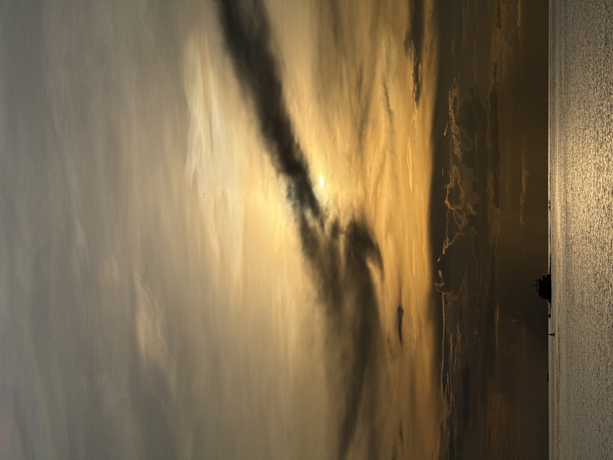 The image depicts a sunset over a calm body of water, likely the ocean, with a beach or shoreline visible on the right. The sky is filled with dramatic clouds, illuminated by the setting sun, creating a gradient of golden and dark hues. The sun itself is partially obscured by dark clouds, casting a warm, diffused light across the scene. The water reflects the sky's colors, shimmering with the sun's rays.
On the far right of the image, along the shoreline, a silhouetted figure of a person can be seen standing near the water's edge, and a darker silhouette of a larger object, possibly a boat or structure, is visible further in the distance. The overall mood is serene and atmospheric, with the vibrant colors of the sunset evoking a sense of peace and tranquility. The location is identified as Koh Phangan, Thailand, suggesting a tropical setting. The time of day is clearly sunset.
