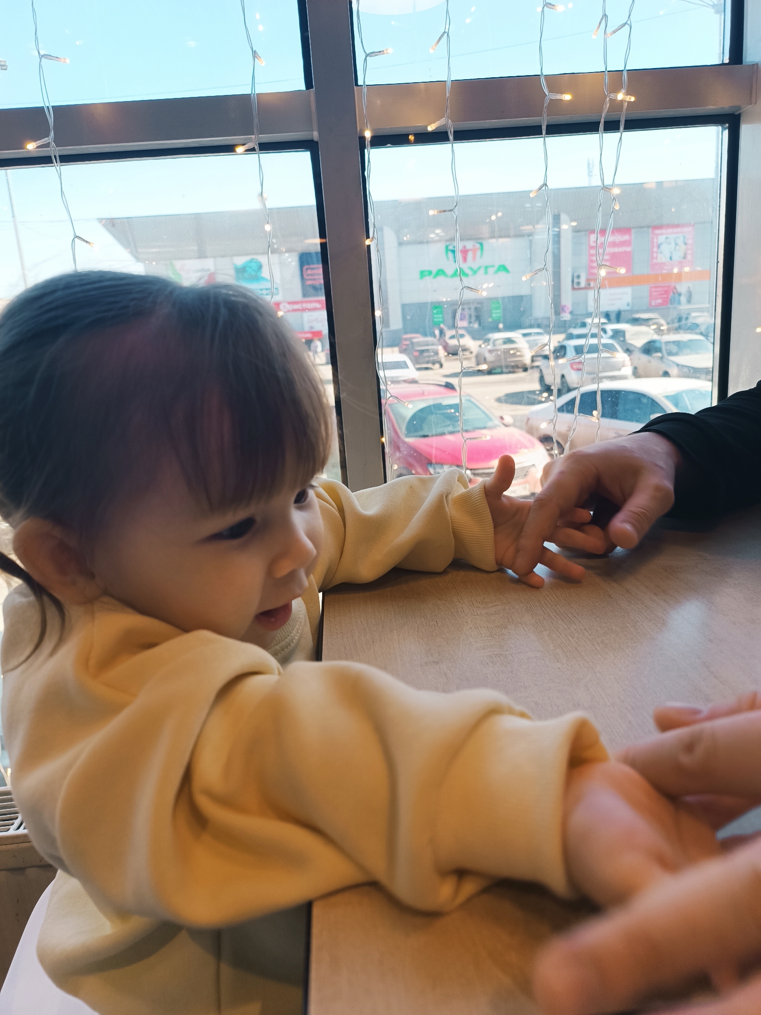 A toddler sits at a table, wearing a light-yellow sweatshirt, with their hands touching the hand of an adult. The child's hair is dark with bangs. Outside the window is a view of a parking lot with cars and a building, identified by a sign as "Радуга", a shopping center in Izhevsk, Russia. Fairy lights hang across the window.