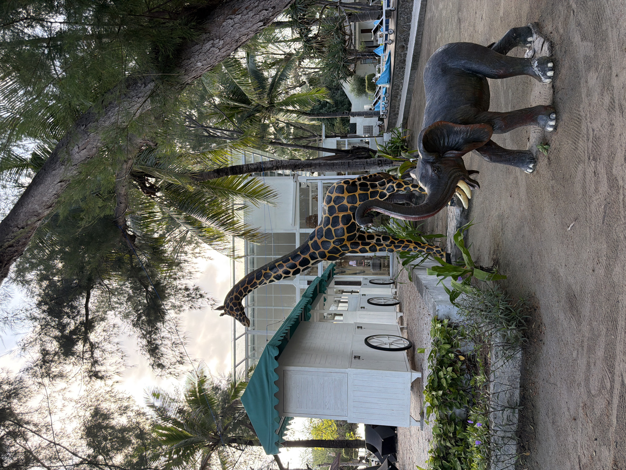 The image depicts an outdoor scene with a playful, somewhat whimsical theme. In the foreground, a life-sized statue of an elephant with its trunk raised stands on sandy ground. Behind the elephant, a large statue of a giraffe with its signature spotted pattern is visible, its head and neck extending upwards.
To the left of the statues is a white, trailer-like structure with a green awning, resembling a food stall or kiosk. It has visible wheels and decorative trim. The structure appears to be set up on a stone foundation. Green foliage and plants are arranged around the foundation of this structure.
In the background, there is a modern building with large glass windows and white architectural elements. Surrounding the area are palm trees and other lush greenery, suggesting a tropical or sub-tropical environment. A swimming pool with lounge chairs and blue inflatable cushions is visible further in the background. The sky above is partly cloudy, with sunlight filtering through the trees. The overall impression is of a resort or a themed recreational area. The location is described as Unknown, Indonesia, but without more information or specific landmarks, the exact city cannot be determined.