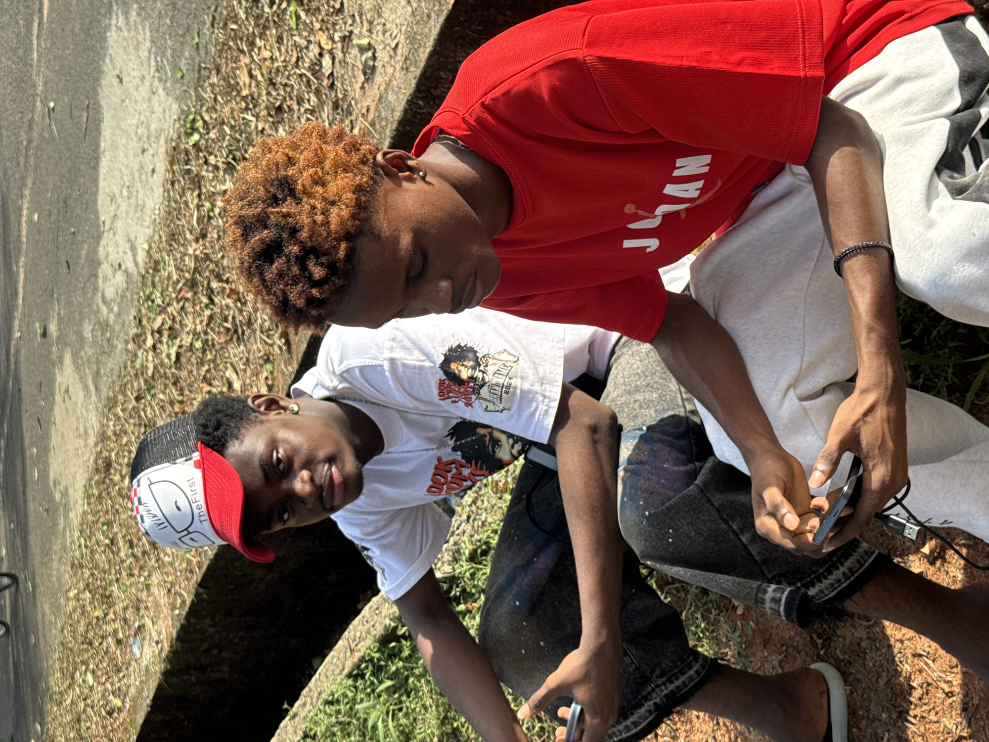 Two dark-skinned young men are outdoors. One man wears a red shirt and light-colored sweatpants with black stripes, and the other wears a white t-shirt with an image on it, a white cap with red trim, and dark jeans. They are both seated on or near a concrete surface and grass, which may be Ihiagwa. The man in red is looking down at a mobile phone. The man in white looks at the camera. A USB cable is lying on the ground next to the man in red.