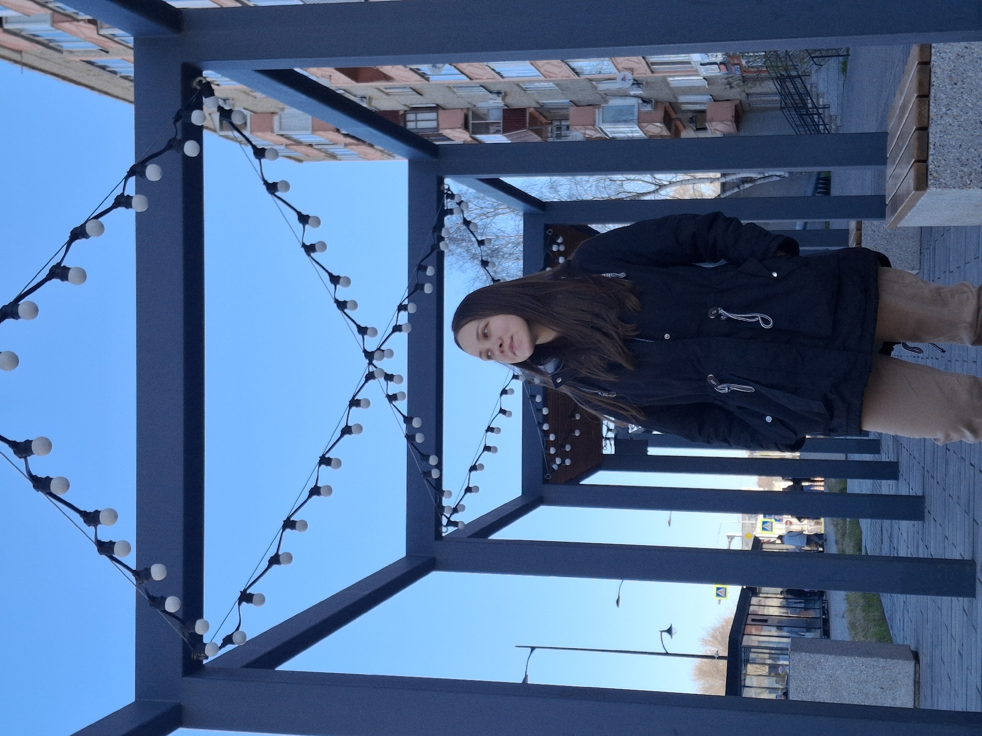 A daytime outdoor scene captured in Sarapul, Russia, shows a young woman standing beneath a modern, dark grey metal pergola adorned with string lights. The image is oriented vertically, with the woman positioned on the right side of the frame, looking slightly upwards and to her right with a gentle, thoughtful expression.
The woman has long, dark brown hair and is dressed in a black winter jacket over what appears to be light brown or khaki trousers. Her hands are in her pockets. The metal structure she stands under features clean lines and connects to a similar framework further in the background, suggesting a walkway or shaded area. Strings of decorative white bulb lights are strung across the horizontal beams of the pergola, creating a festive or inviting atmosphere.
Above and to the left of the woman, a clear, bright blue sky dominates the upper portion of the background. To her right and behind the structure, an urban environment is visible, including a multi-story building with numerous windows, suggesting residential or commercial architecture. Bare trees, indicative of late autumn or early spring, are also scattered in the distance. The ground is paved, and further back, street elements like lampposts and a street sign (a yellow diamond shape, likely a road sign, but no text is discernible) can be seen. A modern-looking concrete bench or planter is also visible on the right.
The overall impression is one of a calm, pleasant day in an urban outdoor space, possibly a pedestrian zone or park. The lighting is bright and even, consistent with daylight.