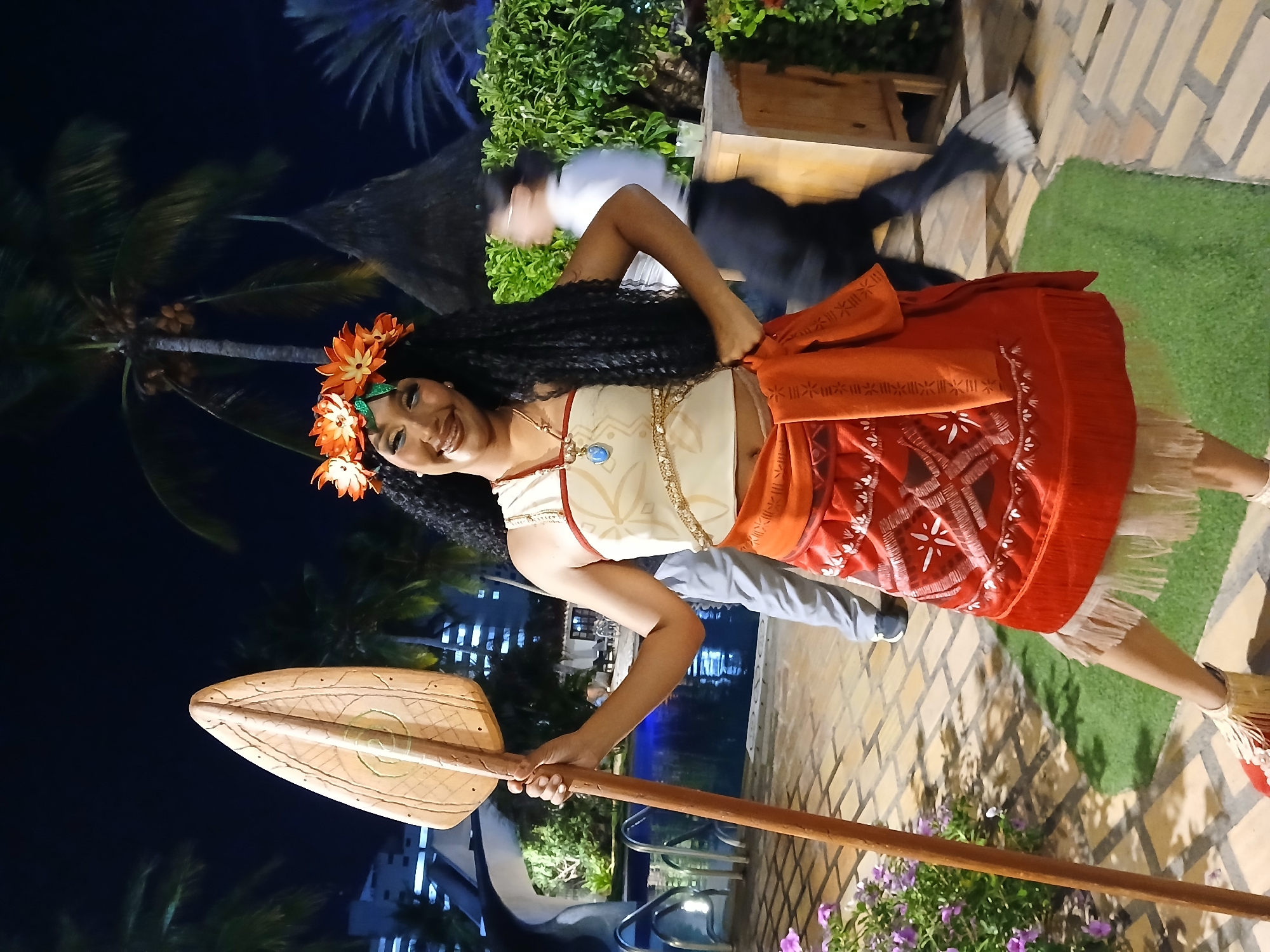 A woman dressed as Moana stands on a brick patio holding a large paddle. She is wearing a floral headpiece and a traditional Moana outfit. Behind her are plants, a palm tree, and a building illuminated at night. The location is Porlamar, Venezuela. There are other people blurred in the background, near a pool and other unidentified objects.