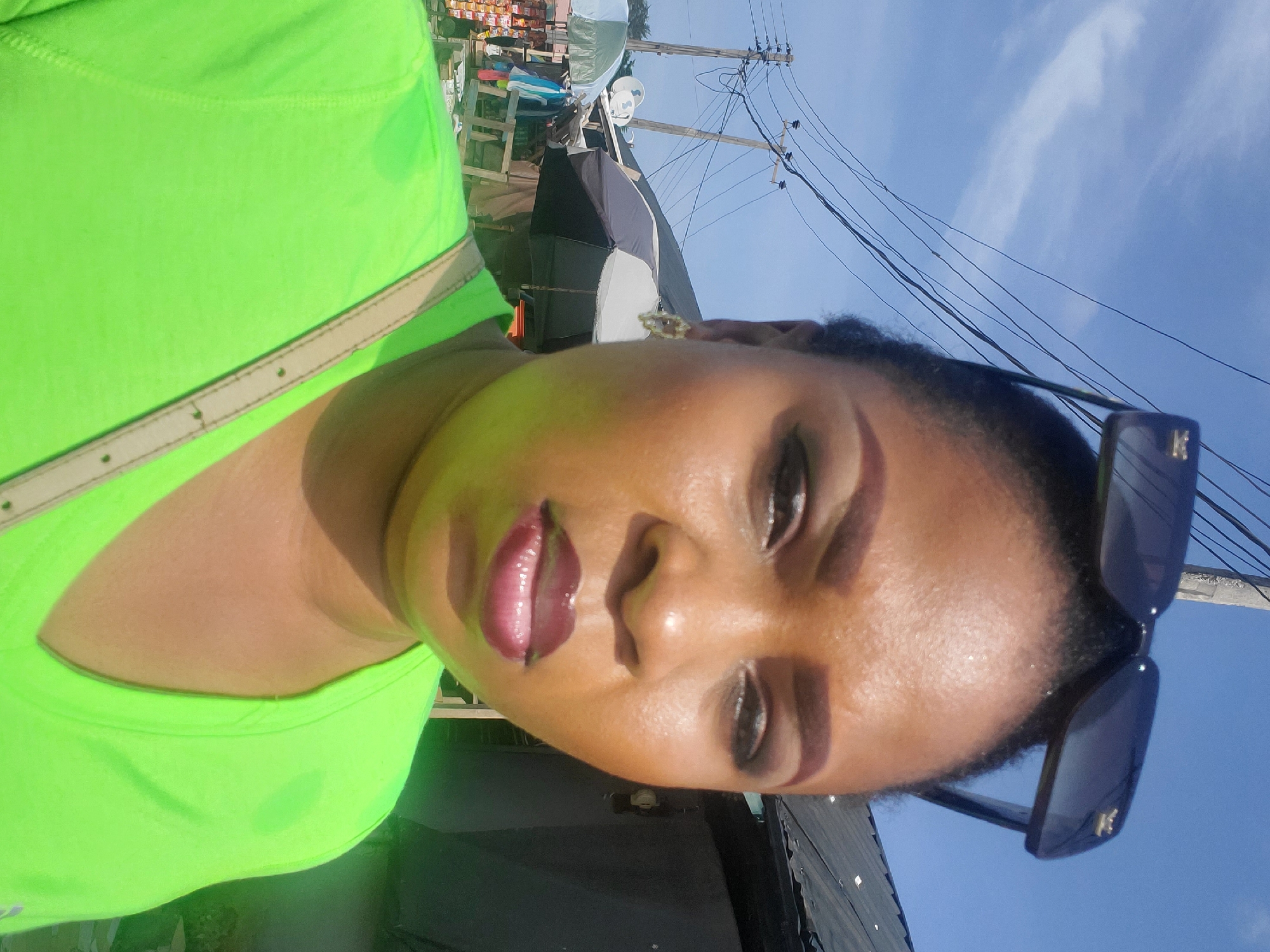 A self-portrait-style image captures a person, presumably female, with medium-dark skin, looking directly at the camera. The subject's face is central and well-lit by bright sunlight, revealing pronounced eye makeup, dark, glossy lips, and a visible sheen on her skin, suggesting warmth or humidity. Dark sunglasses are pushed up onto her head. She is wearing a vibrant neon green top, with a light-colored strap, possibly from a bag, visible across her chest.
The background depicts an outdoor, urban setting under a clear blue sky with sparse white clouds. To the left, behind the subject, are various market stalls or structures, some covered with light-colored tarps or fabrics, and displaying what appear to be goods. On the right, several wooden utility poles are visible, crisscrossed with numerous electrical or communication wires, characteristic of many urban areas in Lagos, Nigeria. The bright, direct sunlight indicates it is daytime, likely mid-day. No explicit text is clearly legible, though a small, unidentifiable emblem is present on the arm of the sunglasses. The overall impression is a casual outdoor moment captured in a bustling environment.