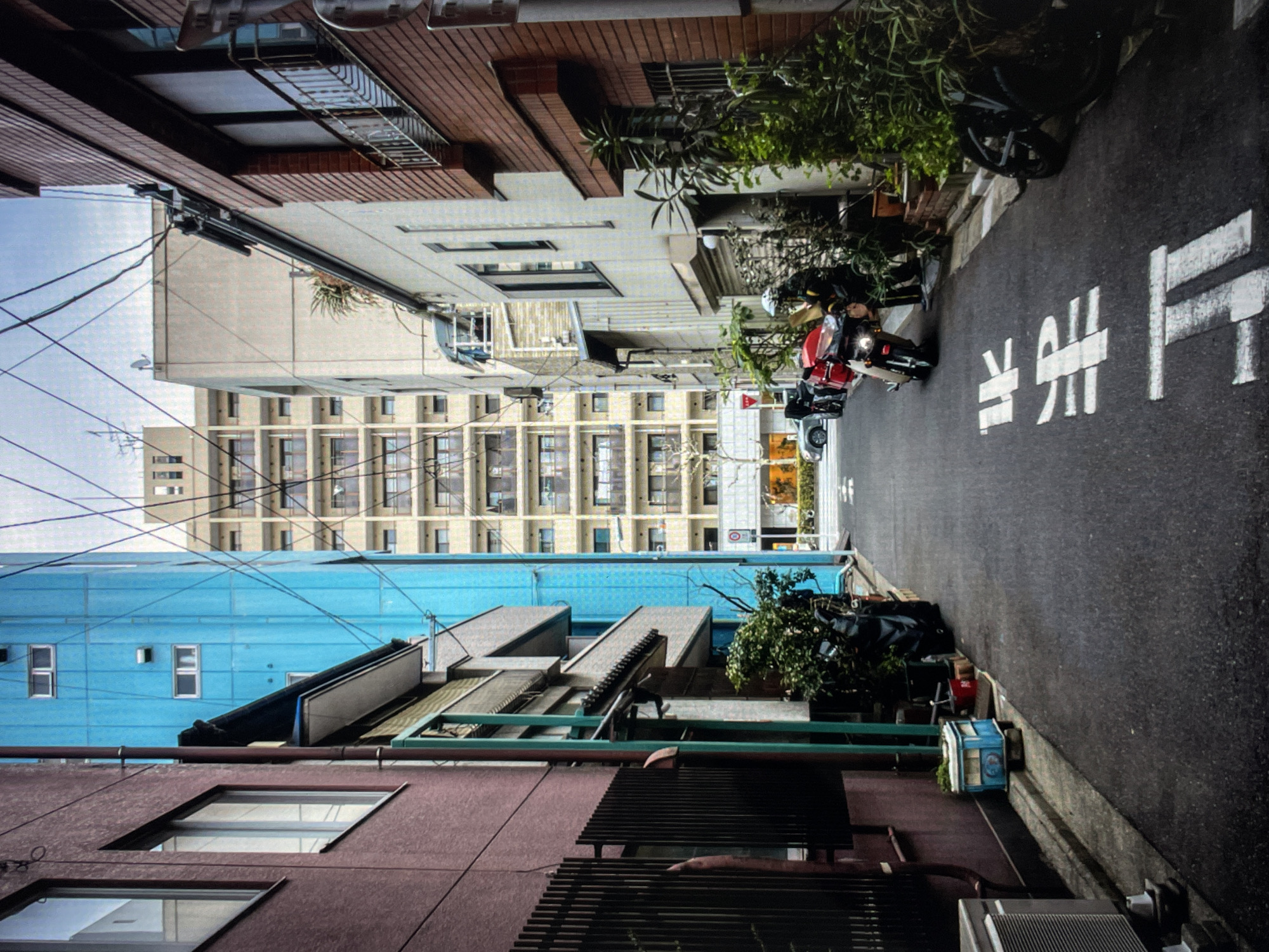 A street view in Unknown, Unknown, shows a narrow road lined with buildings of varying heights and colors. The road surface is dark asphalt with white painted markings, including characters that appear to be Japanese text. Parked motorcycles and other vehicles are visible along the side of the road. The buildings on either side vary in architecture, with some featuring brick, while others have colored facades, such as blue and pink. Above, a tall building with numerous windows rises in the distance, partially obscured by the closer buildings. Power lines crisscross the sky. Plants and vegetation are scattered along the street, adding a touch of greenery to the urban landscape. The image appears to be taken from a perspective that looks straight down the street.