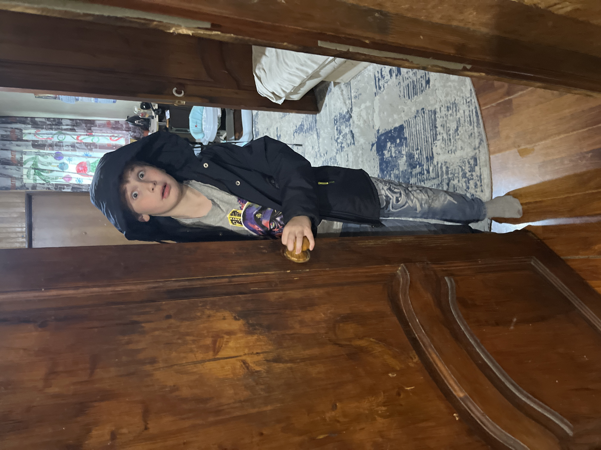 The image, presented in portrait orientation, captures a scene rotated 90 degrees counter-clockwise. A young boy is positioned horizontally across the frame, peeking from behind a dark wooden door. He has light brown hair and is wearing a dark hooded jacket over a gray t-shirt with an indistinct graphic, which features hints of purple and yellow. Blue jeans with a faded pattern and a light gray sock on his left foot complete his attire. His face is turned towards the viewer, with wide, surprised eyes and a slightly open mouth, conveying curiosity or astonishment. His right hand grasps a round, brass-colored doorknob on the sturdy, paneled wooden door, which shows signs of wear and a rich, dark brown finish.
The room visible beyond the door features a blue and white patterned rug on what appears to be a light-colored wooden floor. To the left, a window is partly obscured by light-colored curtains adorned with colorful, abstract shapes that resemble balloons or festive decorations. In the deeper background, a white sheet or blanket drapes over unseen furniture, and a white plastic container is dimly visible. The domestic setting suggests a home in Vesyoloye, Russia. No discernible text is legible on the boy's t-shirt graphic.