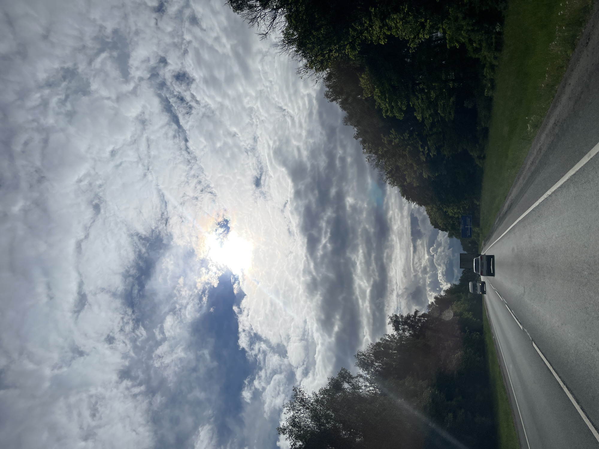 The image shows a view from a vehicle driving on a highway, potentially near Ol'khovka, Russia, but the specific location cannot be confirmed. The sky is filled with predominantly white and grey clouds, with the sun partially visible through a break in the cloud cover. The roadway is asphalt and has a white lane divider. Two vehicles are visible ahead, seemingly traveling in the same direction. The roadside is lined with trees. The image is oriented with the sky on the left and the road on the right.