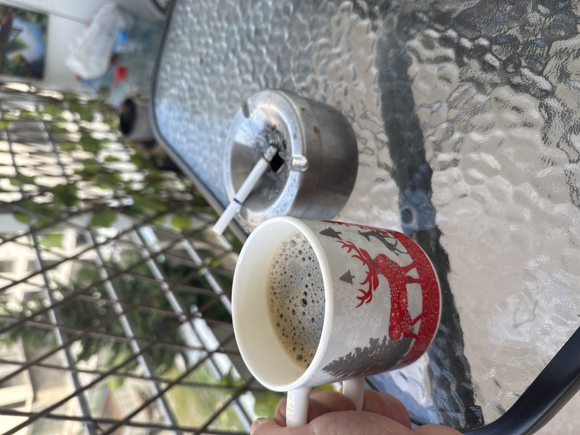 A close-up shot focuses on a white mug filled with dark coffee, adorned with a festive red reindeer and snowflake pattern. The mug is held by a hand, with the index finger and thumb gripping the handle. To the left of the mug, an ashtray containing a lit cigarette is visible. The ashtray appears to be made of metal and has some ash and soot. Both the mug and ashtray rest on a textured glass tabletop. In the background, through a metal mesh fence, lush green foliage and buildings can be seen, suggesting an outdoor setting. The lighting implies it is daytime. The scene evokes a sense of casual relaxation, possibly a morning coffee break or an evening smoke on a balcony or patio.